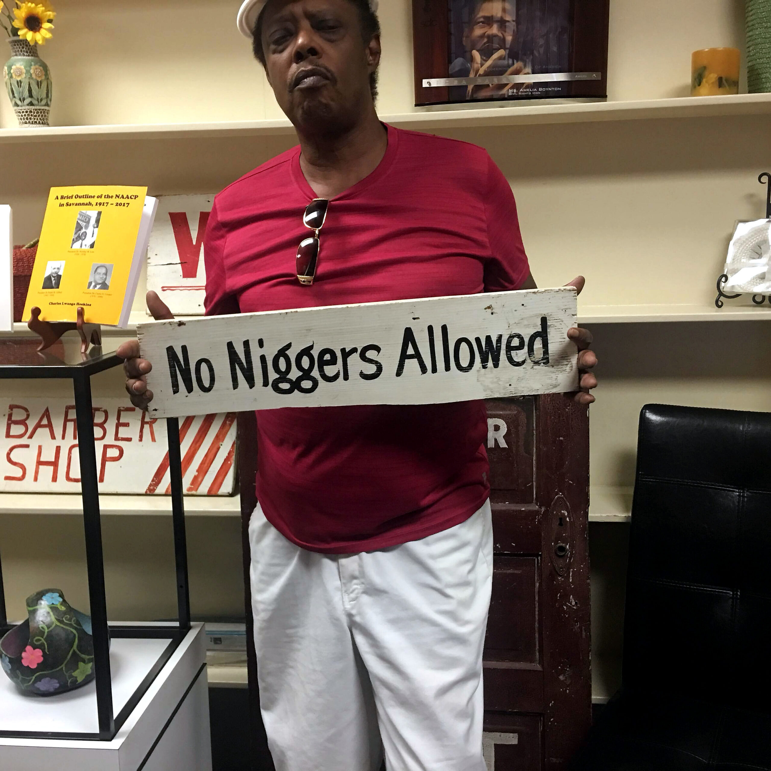 Man in a red shirt holding a weathered sign that reads 'No Niggers Allowed' in a shop setting.