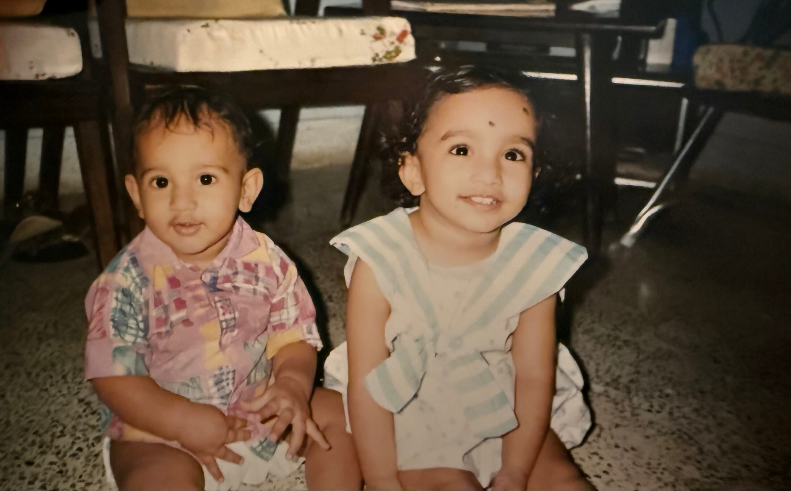 Two young children sit on a speckled floor under a dining table, smiling at the camera with colorful outfits.