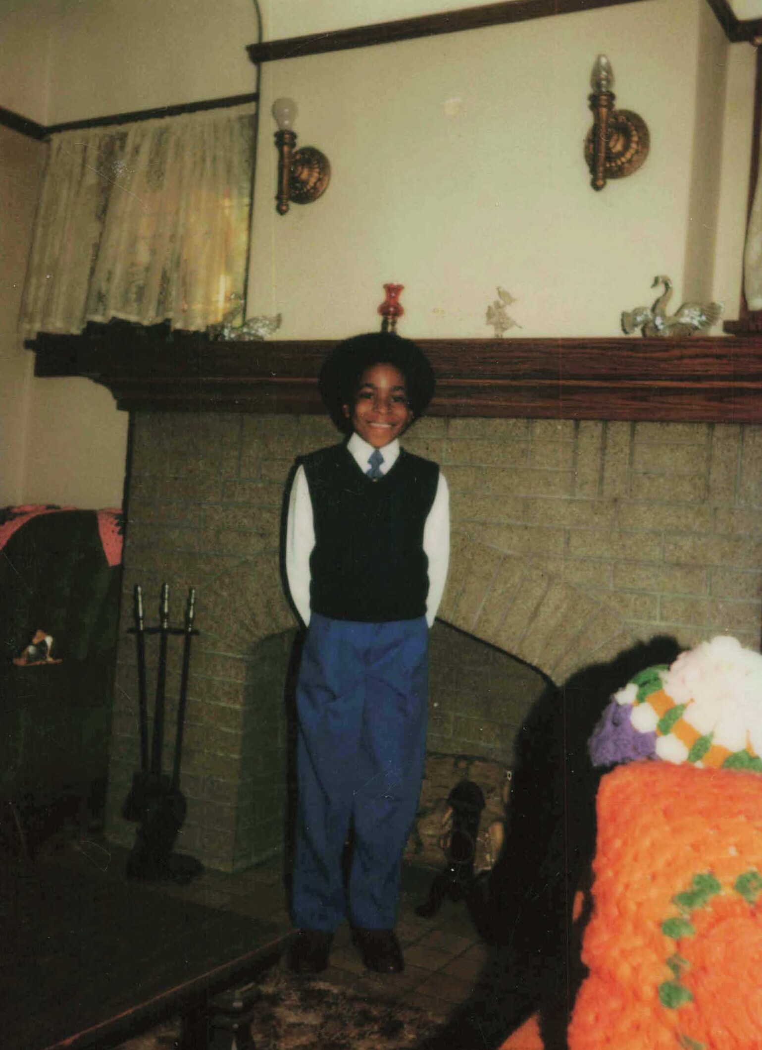 Smiling boy with an afro in a vest and tie, standing in a living room in front of a brick fireplace mantle with decorations.