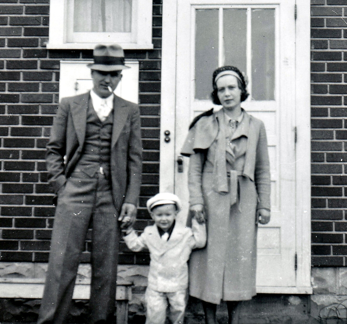 Vintage photo of a family of three standing outside a brick house: man in a suit and hat smoking, woman in a coat with a headscarf, and a young child between them holding their hands.