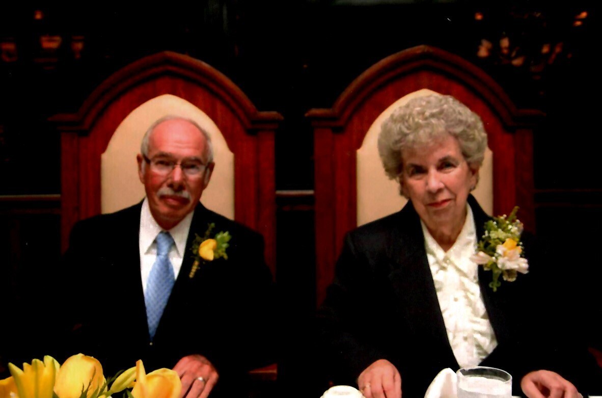 Elderly couple in formal attire seated at a banquet table with yellow flowers in front.