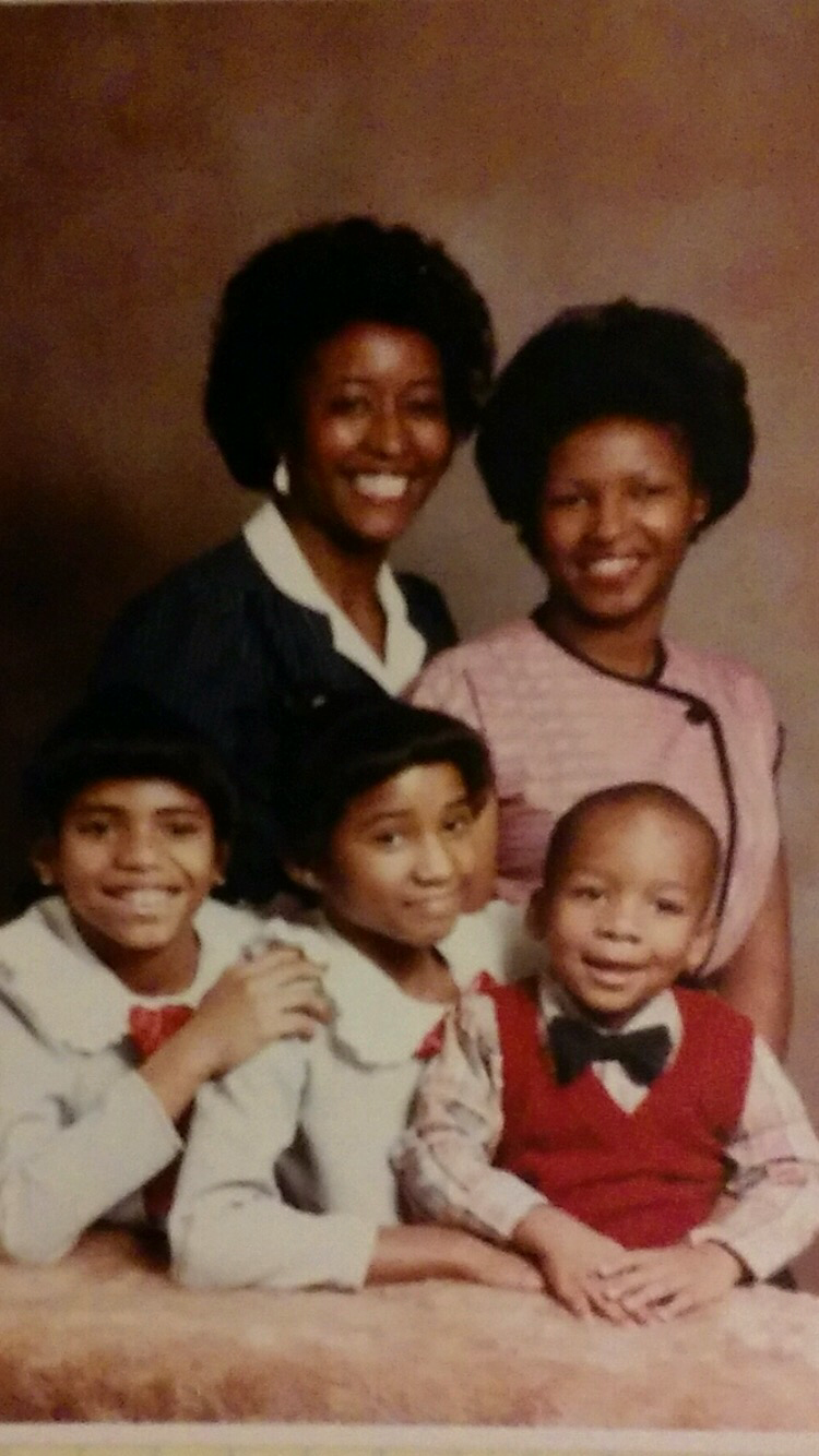 A family portrait of five posing in a studio: two adults behind three children, all smiling at the camera.