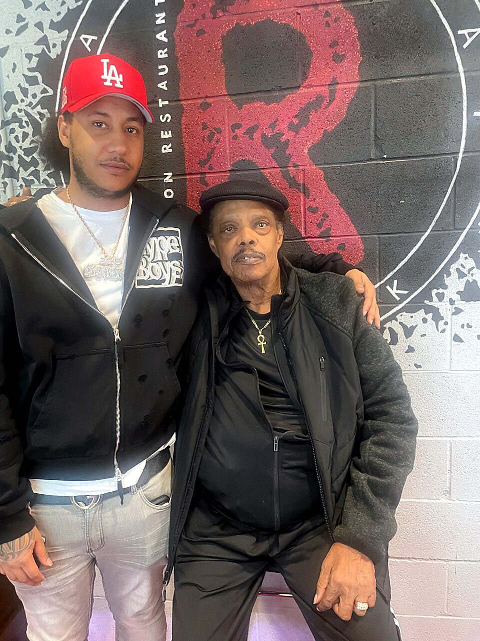 Two men pose in front of a mural: a younger man wearing a red LA cap and black jacket, and an older man in a black jacket and hat with an ankh necklace.