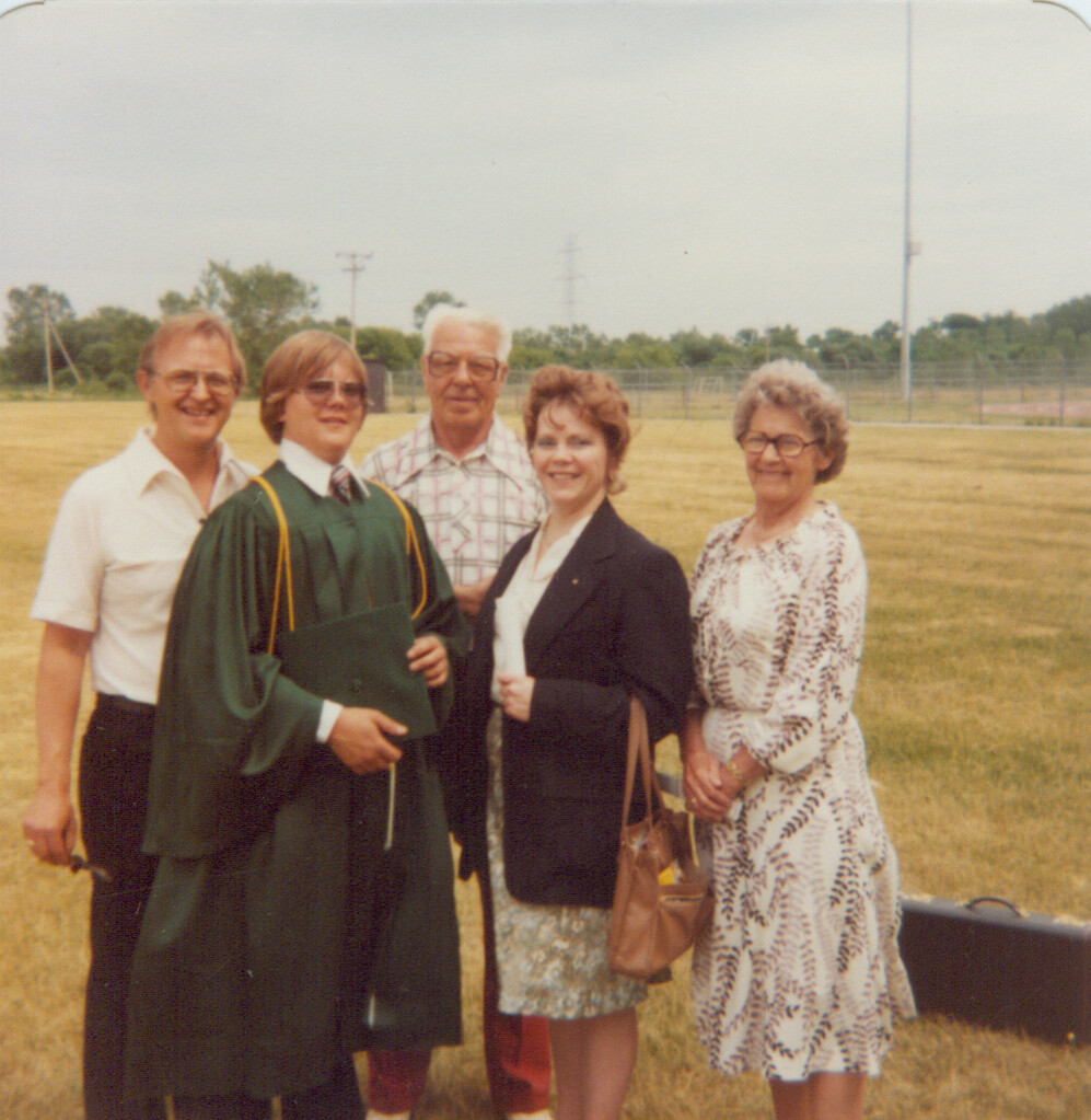 Graduate in green cap and gown holding a diploma poses with four family members outdoors on a sunny, grassy field.