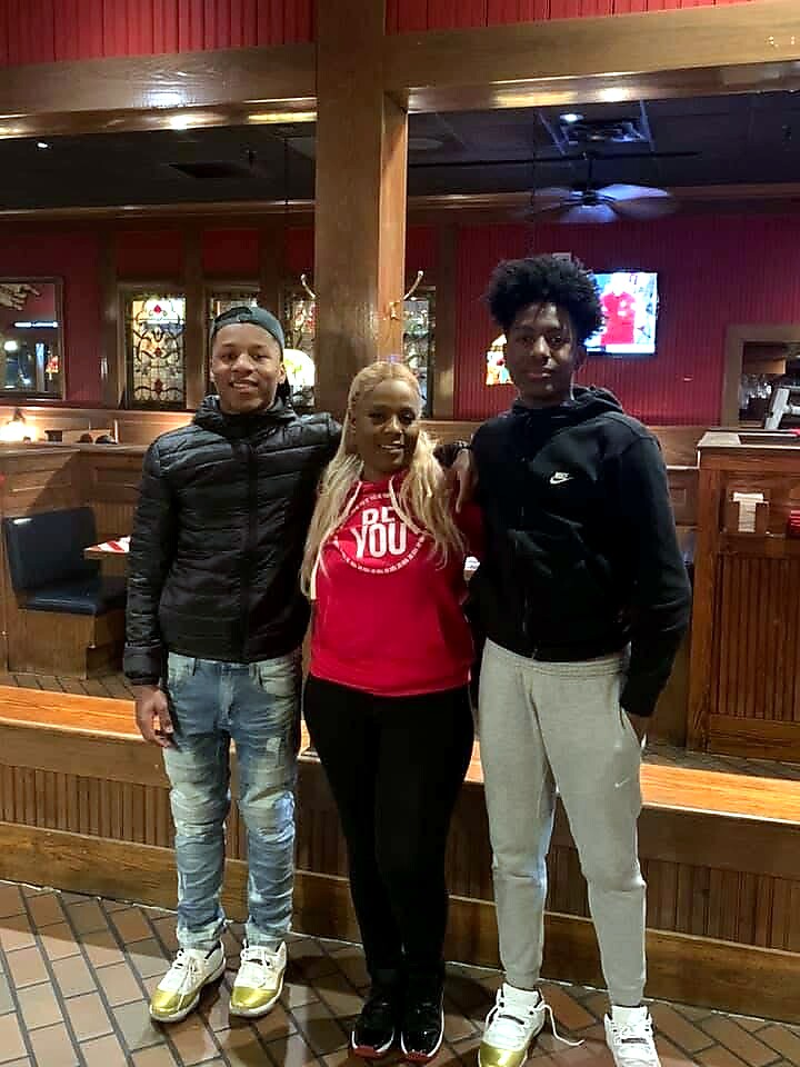 Three friends pose for a photo inside a wood-paneled restaurant, smiling at the camera. The person in the middle wears a red hoodie; the two on the sides wear dark jackets.