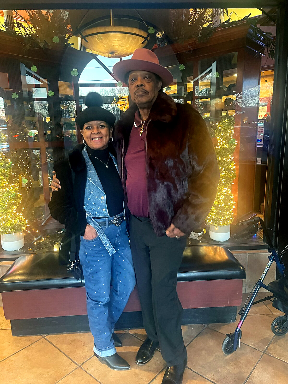 Smiling elderly couple posing outside a storefront with festive lights; woman wears a black hat and denim vest, man wears a pink hat and fur coat.