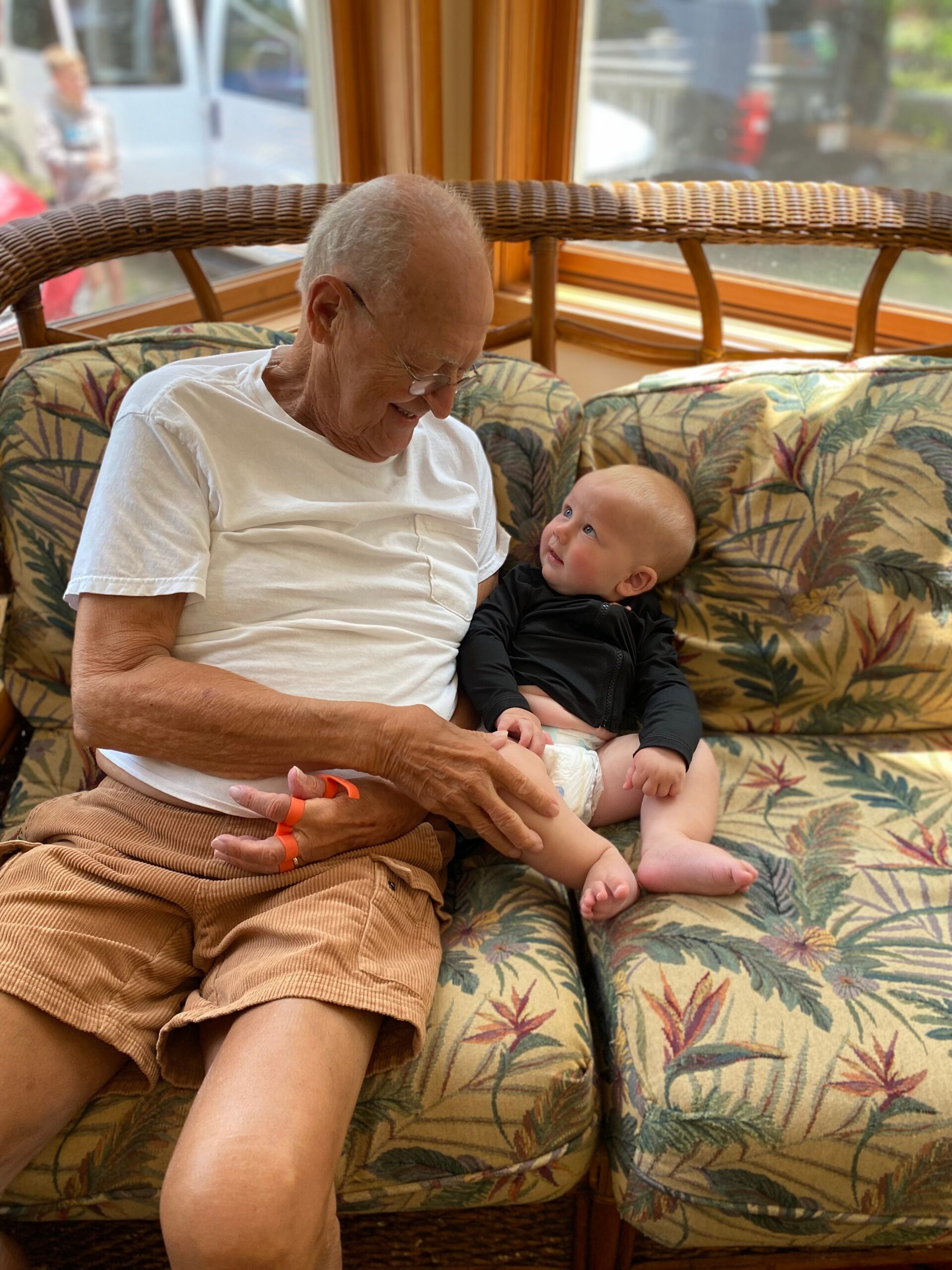 Grandfather in a white shirt sitting on a floral wicker couch, smiling at a baby resting on his lap who looks up at him in a bright room.