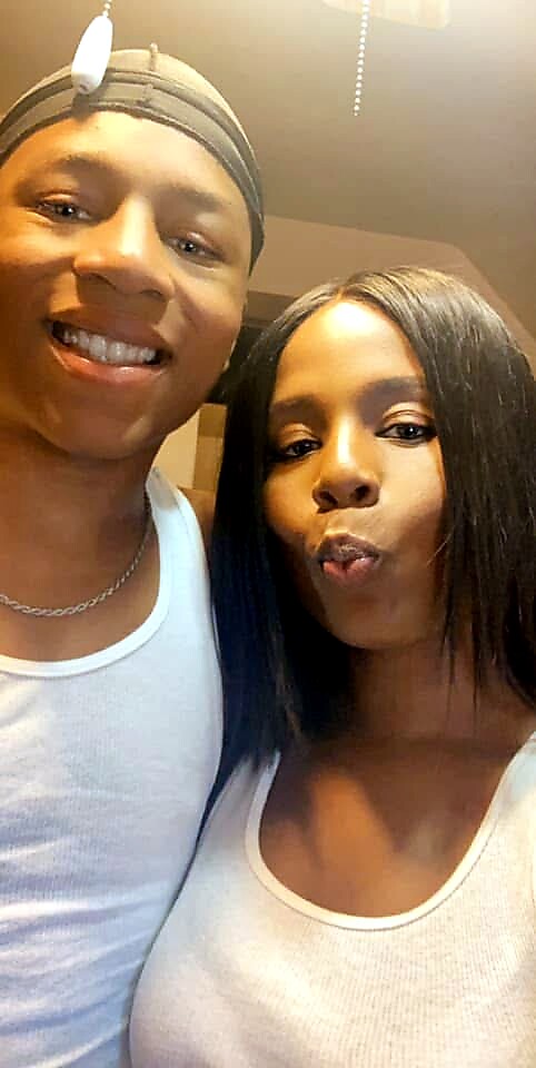Two friends posing for a selfie indoors; man on the left wears a durag and white tank top and smiles, while the woman on the right with straight hair makes a playful pout.