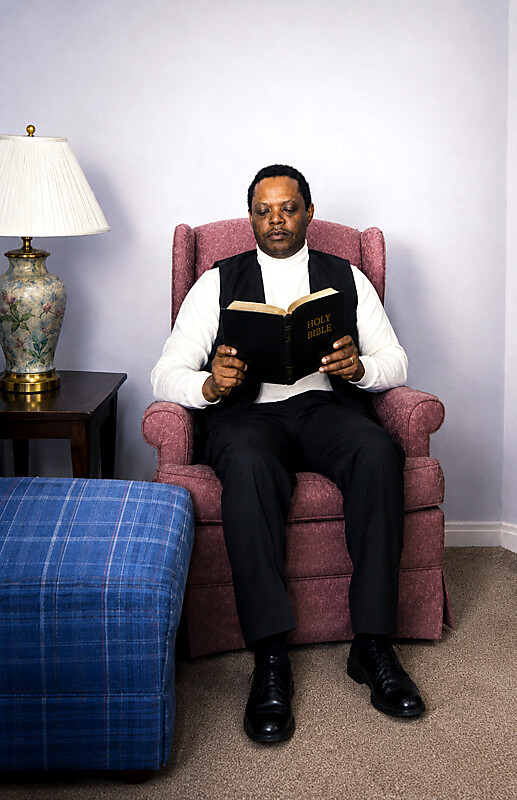 A man in a white turtleneck and black vest sits in a pink armchair, reading a Holy Bible.