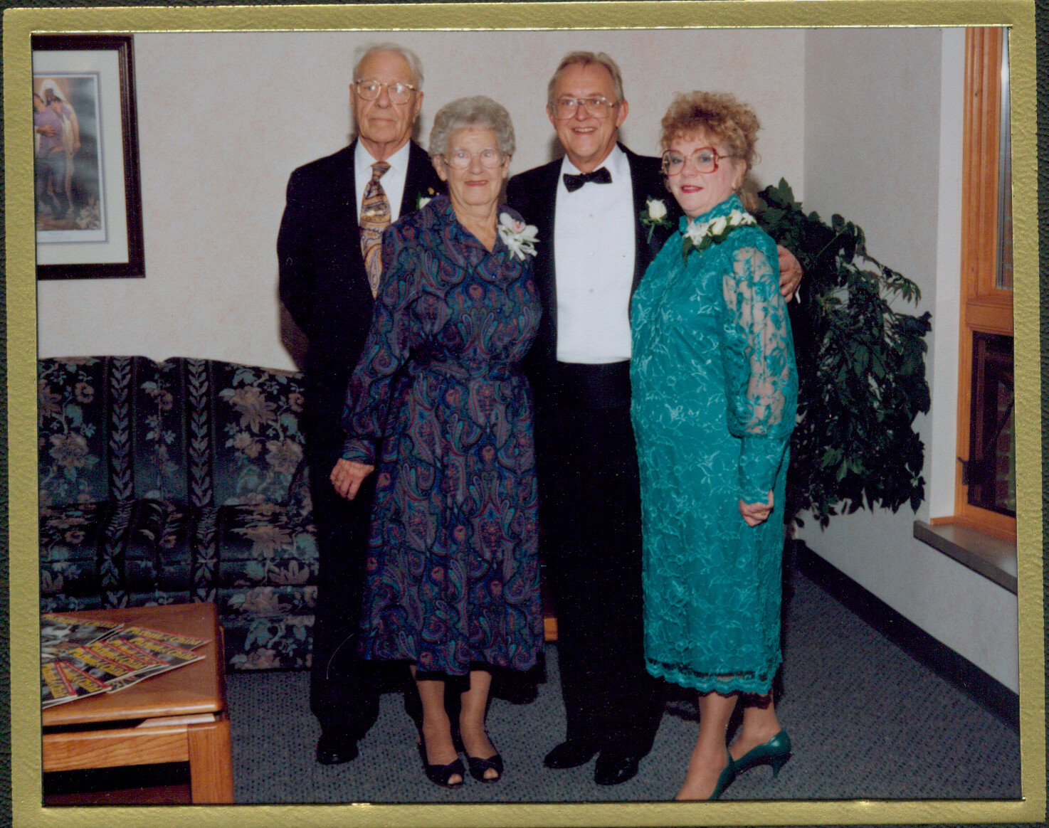 Four older adults posing for a formal photo in a living room: two men in suits (one with bow tie) and two women in colorful dresses wearing corsages.