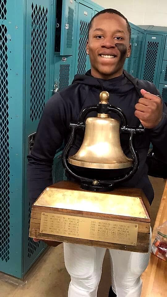 Smiling young man in a locker room holds a large brass trophy bell mounted on a wooden base with an inscription plaque.