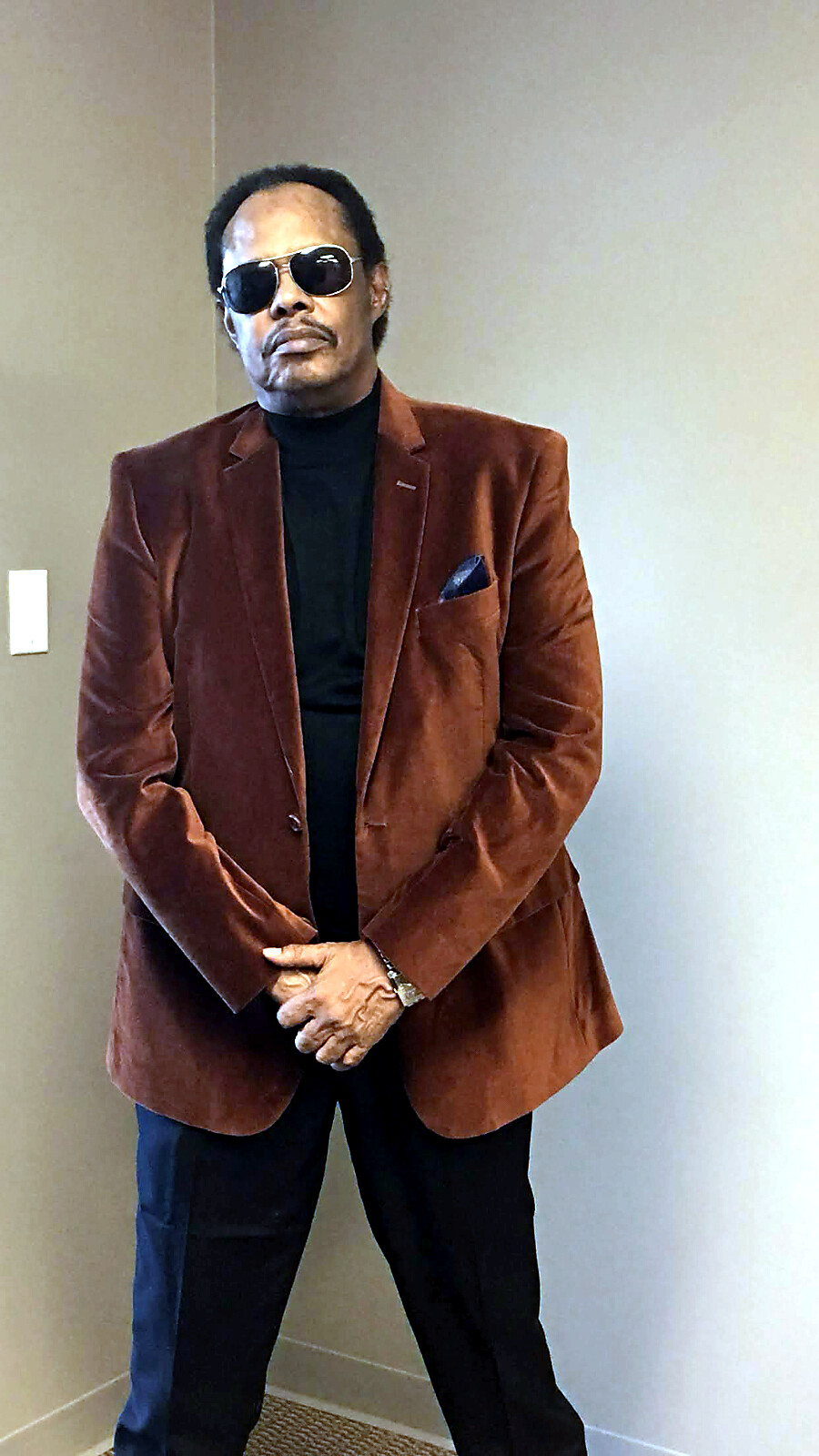 A man in a brown velvet blazer, black turtleneck, sunglasses, and a blue pocket square stands with hands clasped indoors.