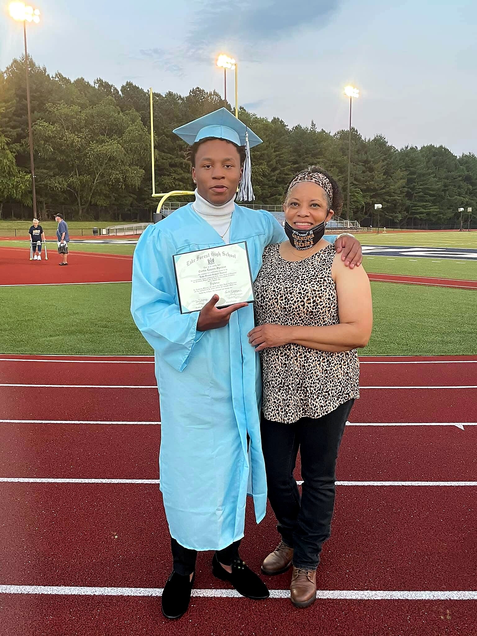 Graduate in a blue cap and gown holding a diploma, posing with a smiling woman on a track field.