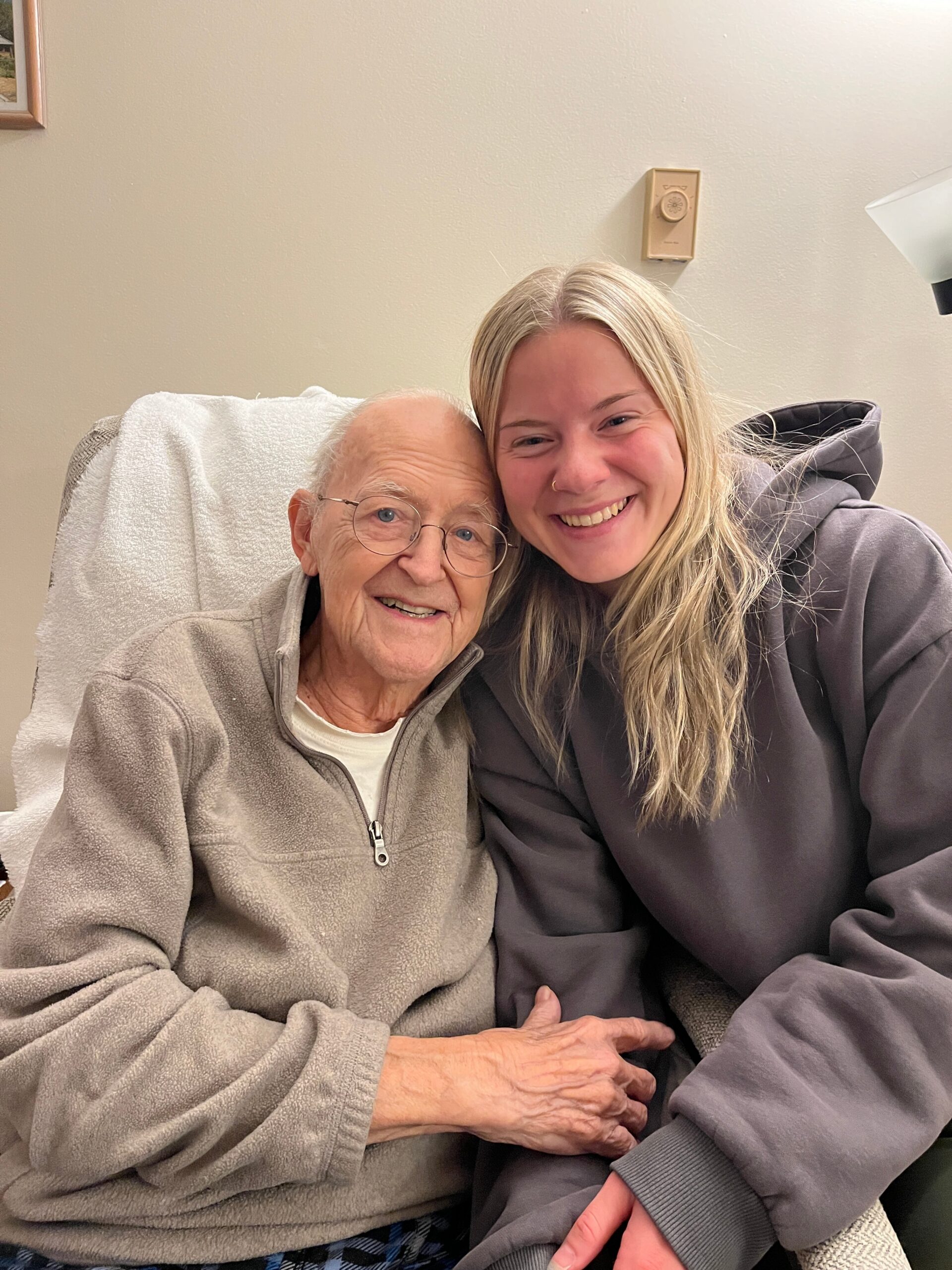 Two women hug and smile at the camera, an elderly woman with glasses on the left and a younger woman on the right wearing gray clothing.
