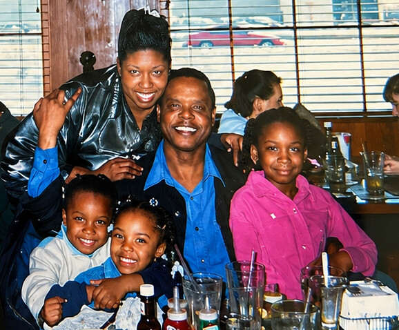 Family of six posing together at a restaurant, smiling for the camera with drinks and condiments on the table.