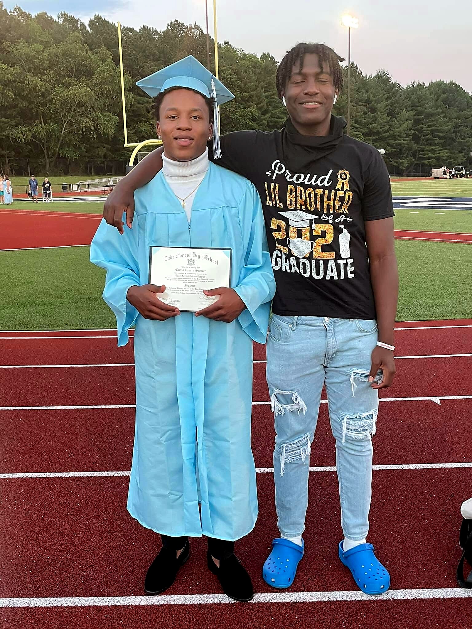 Two young men on a football field; one in a blue cap and gown holds a diploma, the other in a black shirt that says 'Proud Lil Brother 2021 Graduate'.