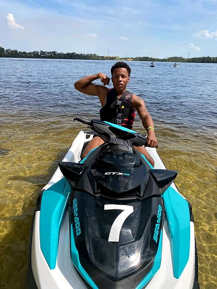 Young man in a life jacket poses on a turquoise-and-black Sea-Doo jet ski in shallow water by the shore, flexing his bicep at the camera.