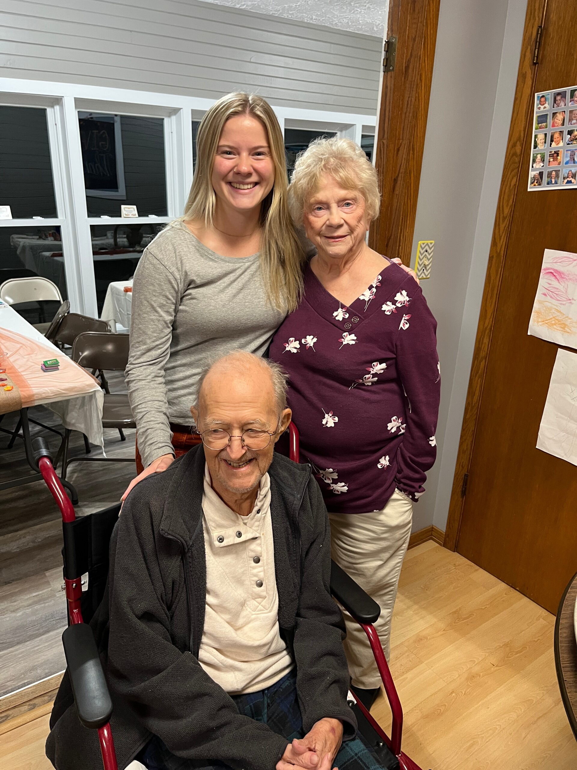 Three generations posing for a photo indoors: an elderly man in a wheelchair in front, and two women smiling behind him.