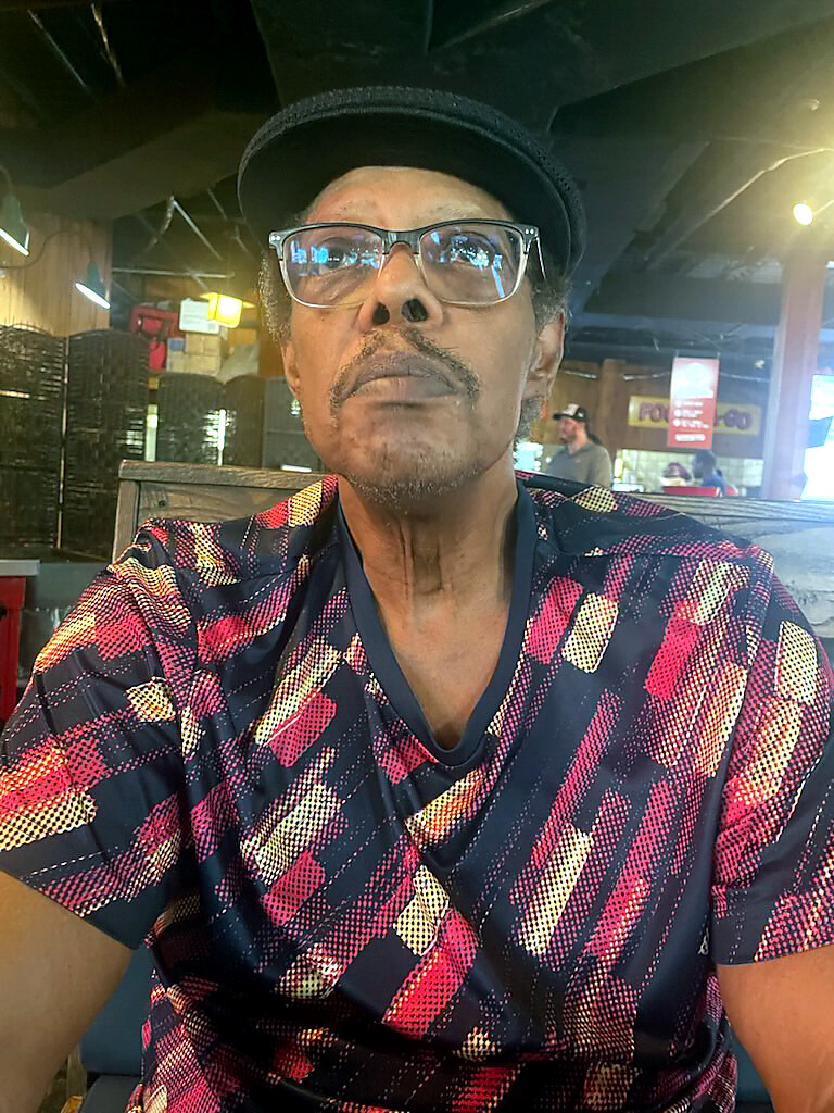 Man wearing a black hat and glasses, in a colorful plaid shirt, sitting in a cafe and looking at the camera.
