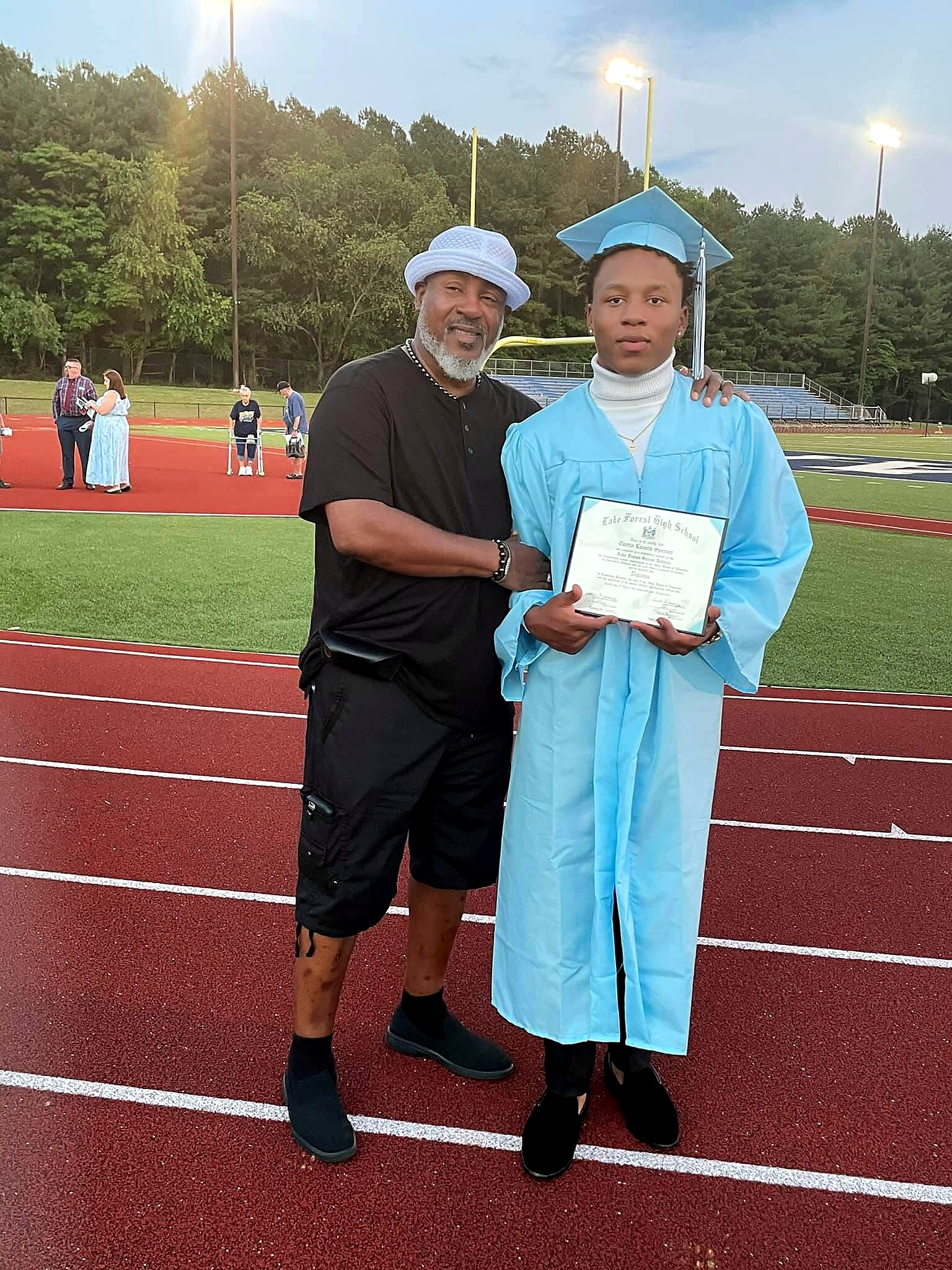 Man in a black shirt and white hat posing with a graduate in a blue cap and gown on a track field, holding a diploma.