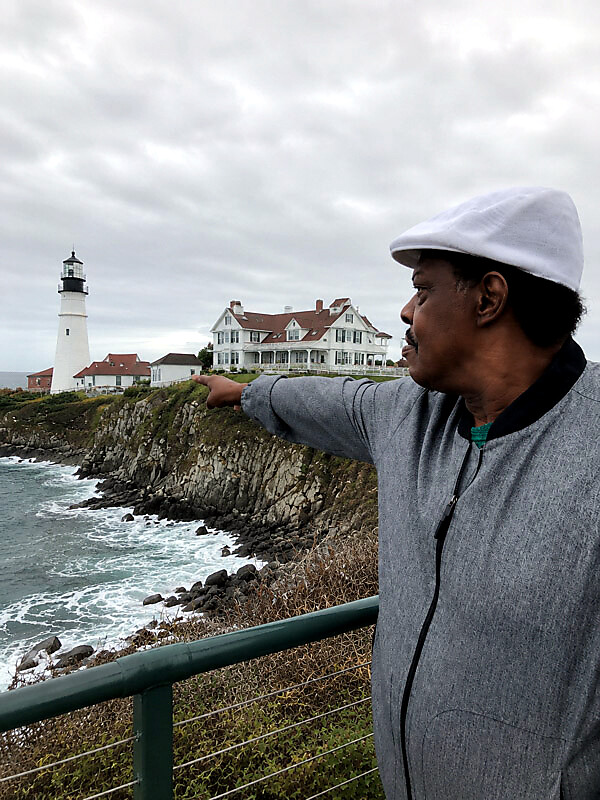 Man in a gray jacket and white cap points toward a lighthouse on a rocky coastline with sea spray and houses in the background.