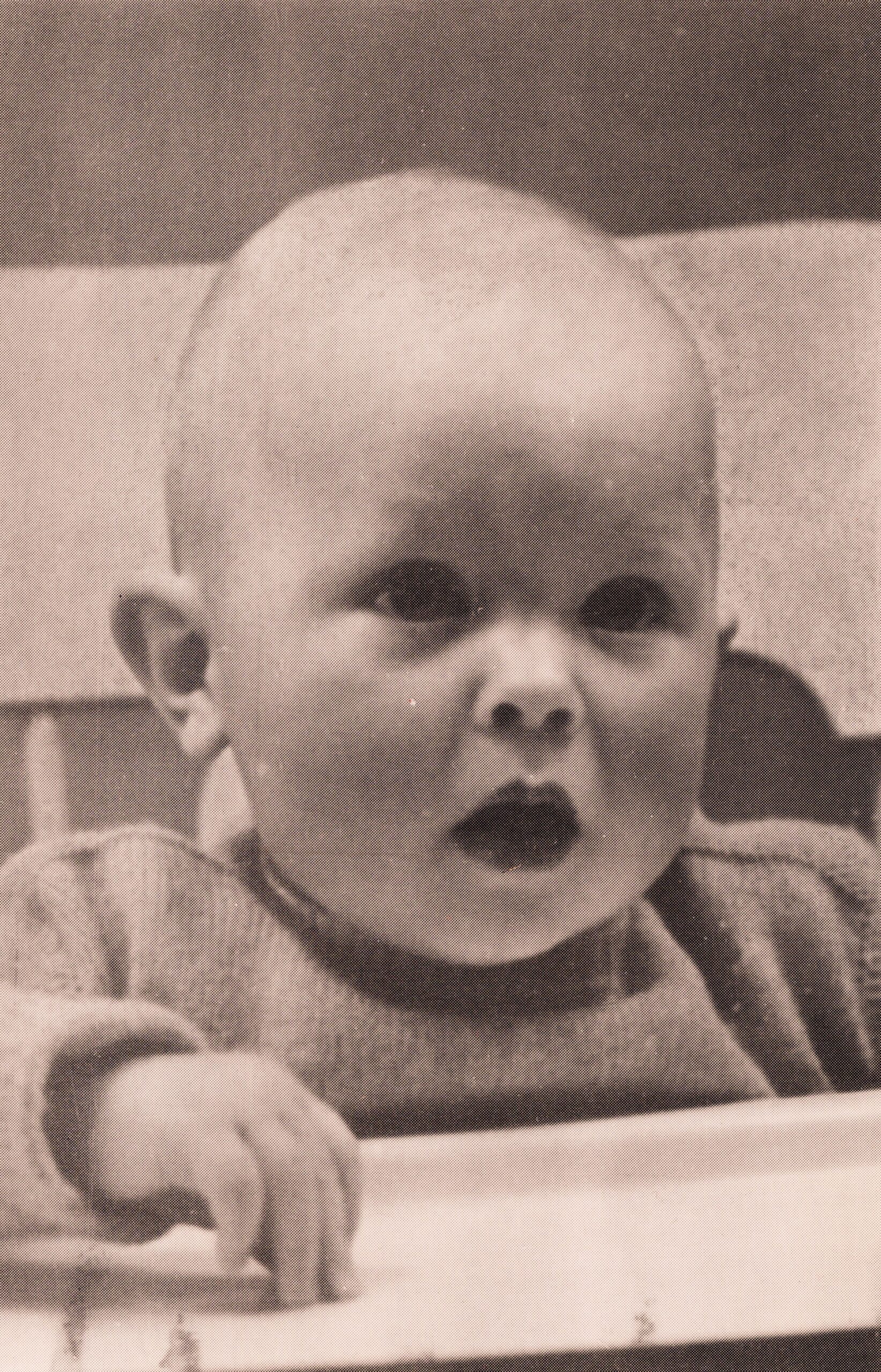 Close-up of a baby with an open mouth, seated at a table and reaching toward a plate in front of him.