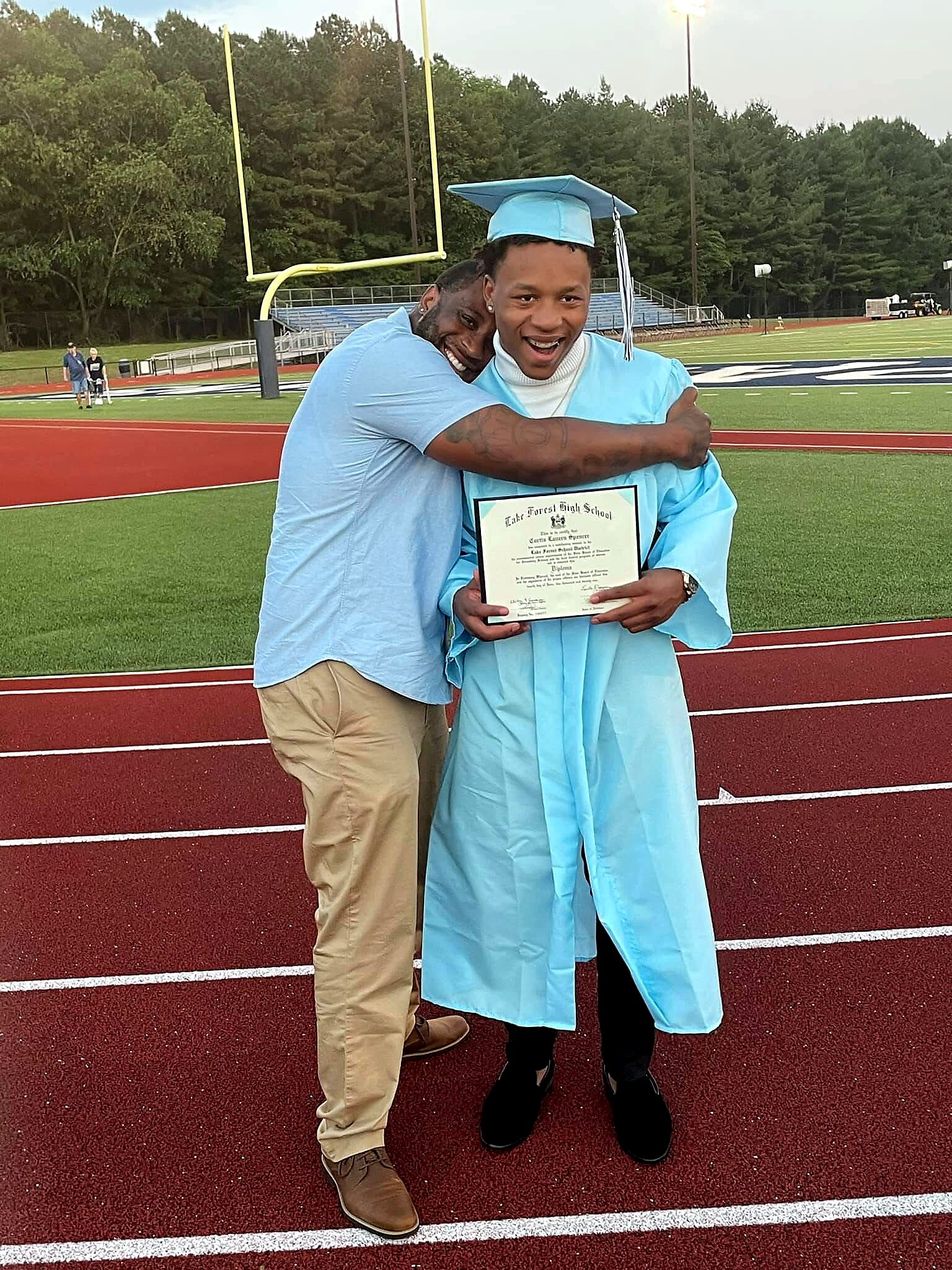 Graduate in a light blue cap and gown smiles while holding a diploma on a track field, hugged by a man in a blue shirt nearby.