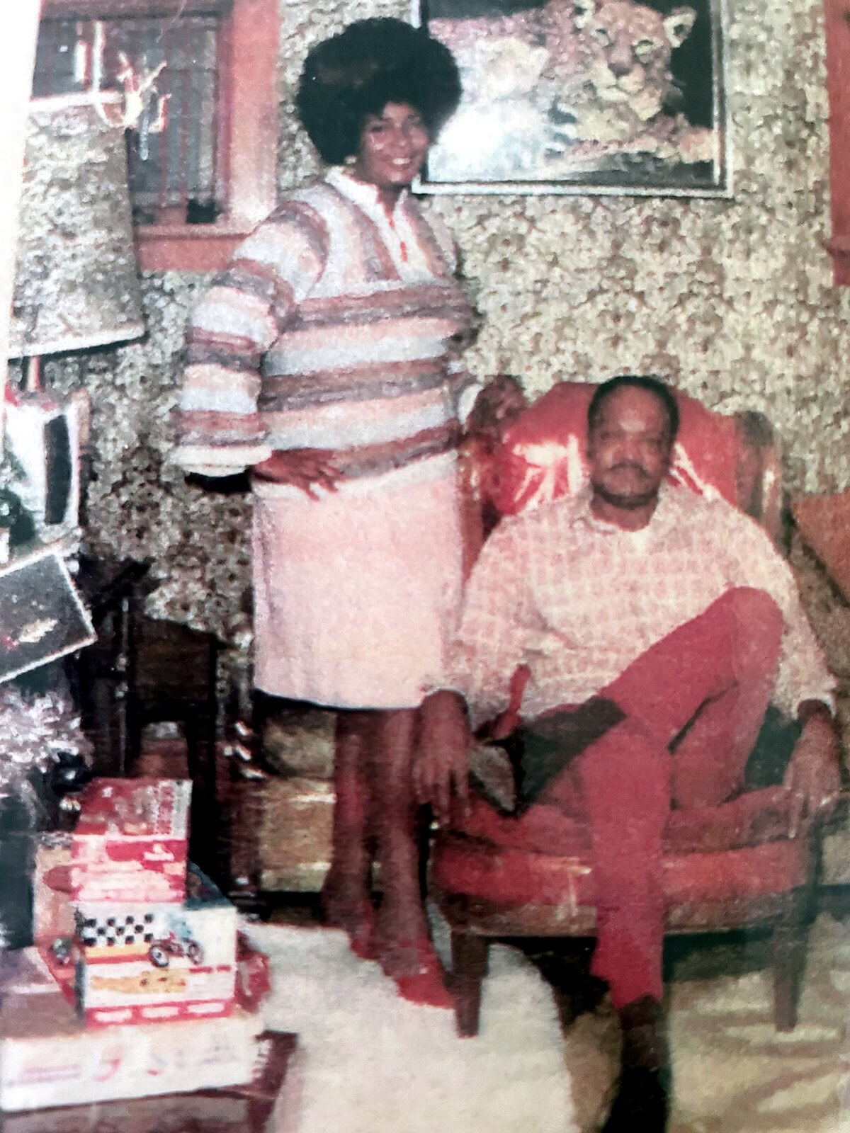 An older woman stands beside a seated man in a cozy, cluttered living room with textured wallpaper and framed art behind them.