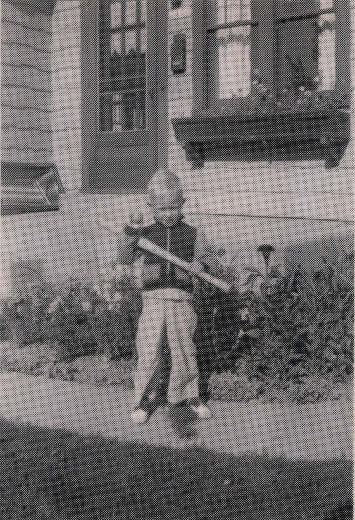 A young child stands in a front yard, holding a long rifle and waving, with a house and garden behind them.
