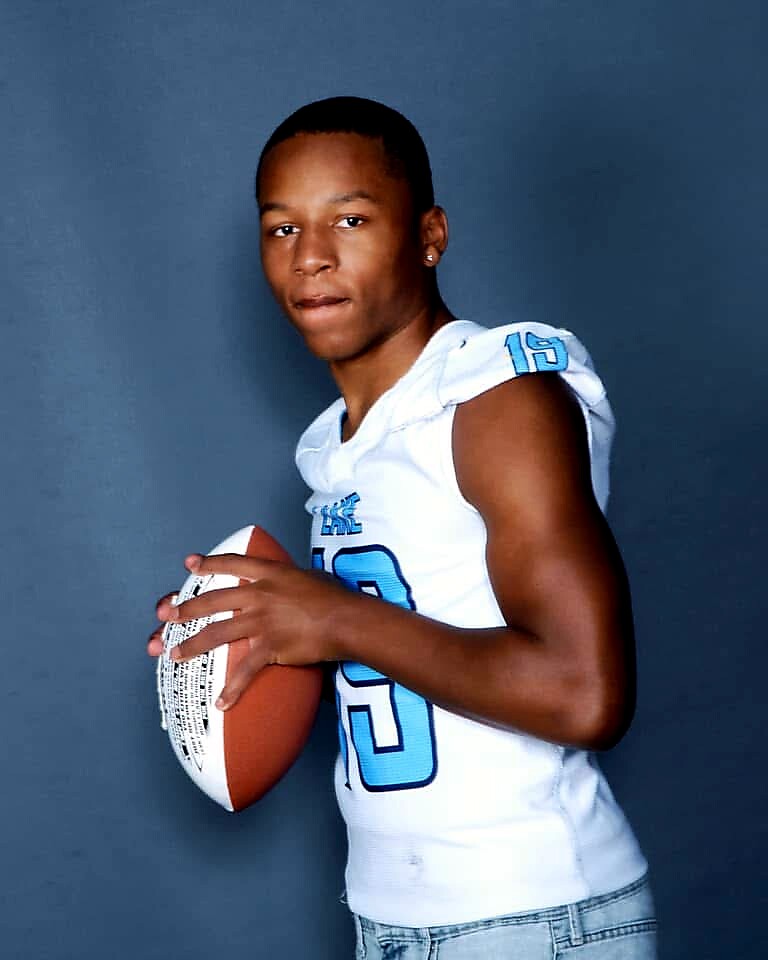 Young man in a white football jersey holding a football, facing camera against a gray backdrop, number 15 visible on sleeve.