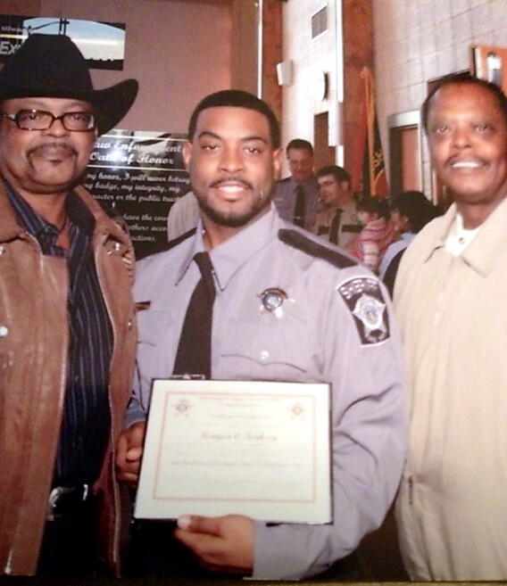 Three men pose indoors; center man in a police uniform holds a certificate, flanked by a man in a cowboy hat and another in a beige shirt.