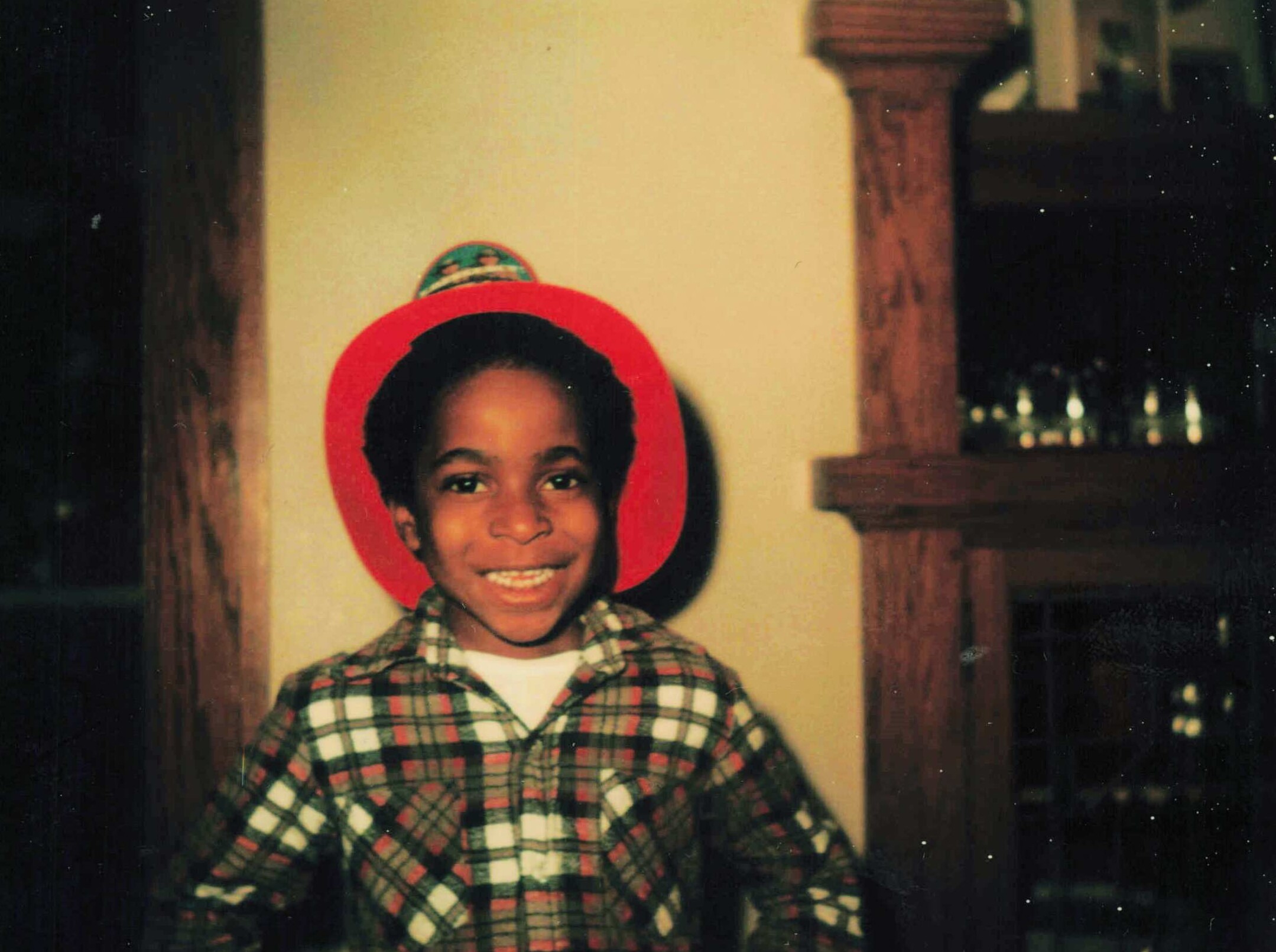 Smiling child wearing a bright red circular hat/hood and a plaid shirt, standing indoors in front of a wall.