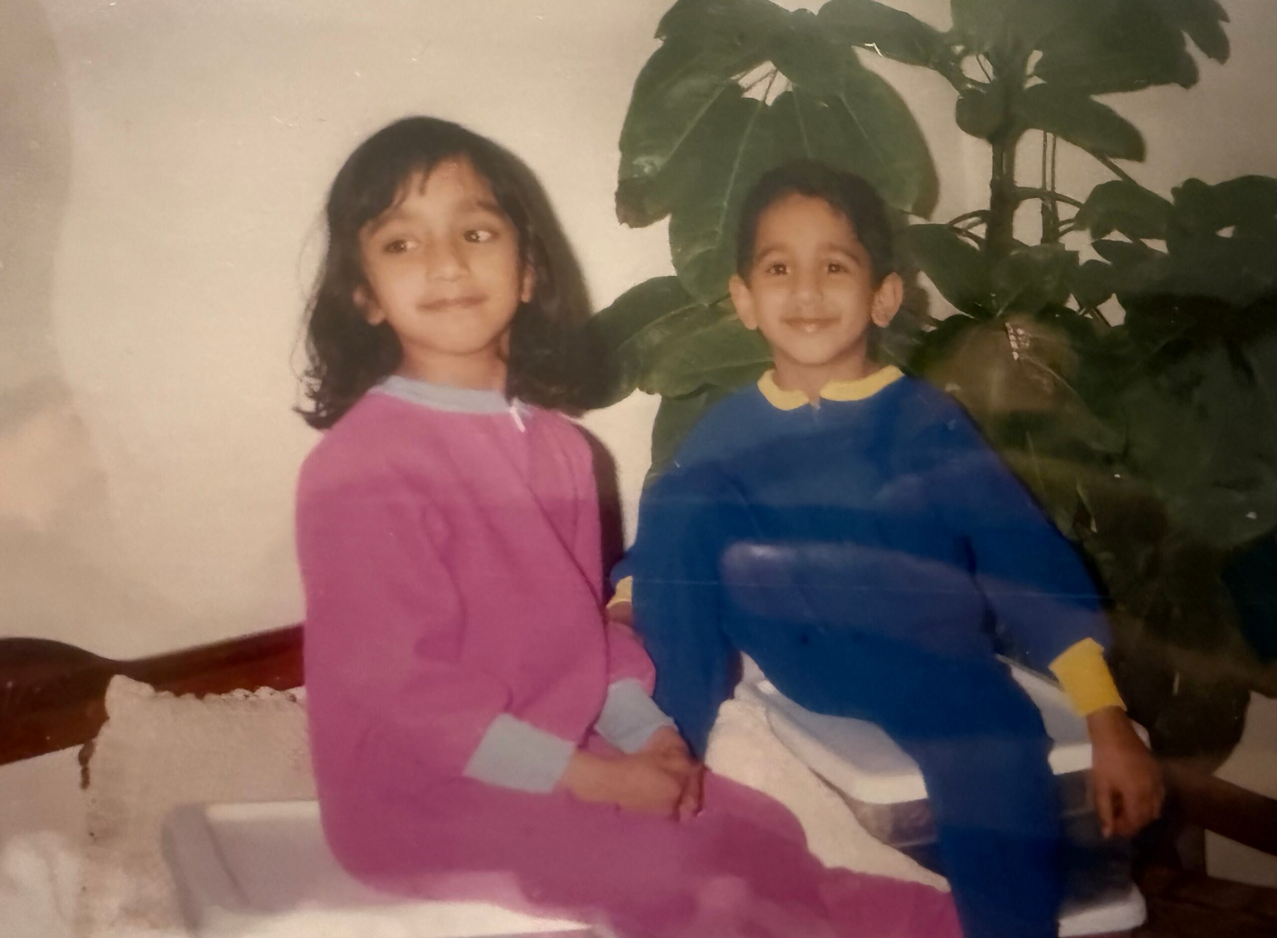 Two young children standing indoors beside a large leafy green plant; girl in pink on the left and boy in blue on the right, both smiling.