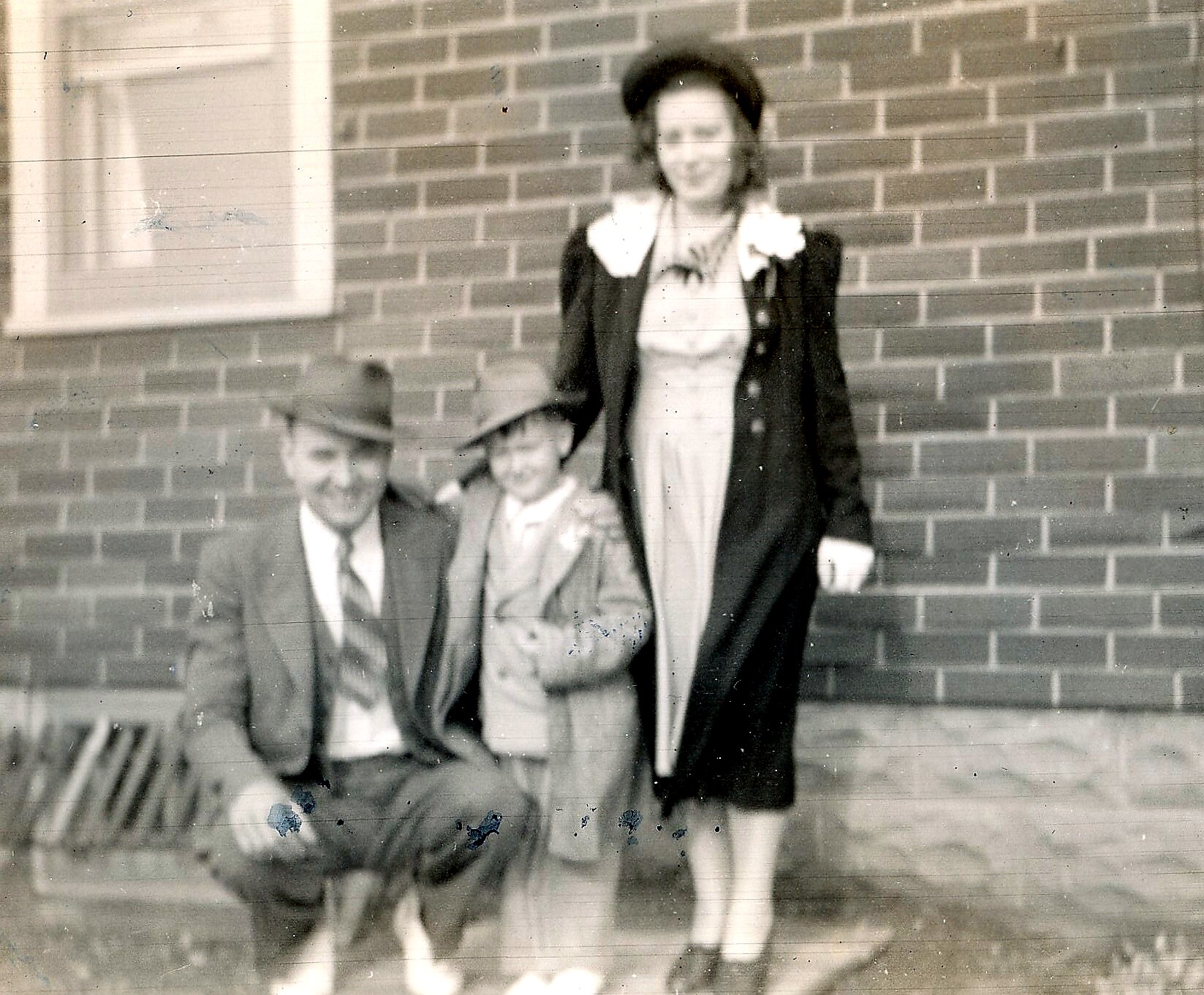 Three costumed people posing outside against a brick wall: a man seated left in a suit and hat, a child sitting center, and a woman standing right in a dress and coat with large white collar.