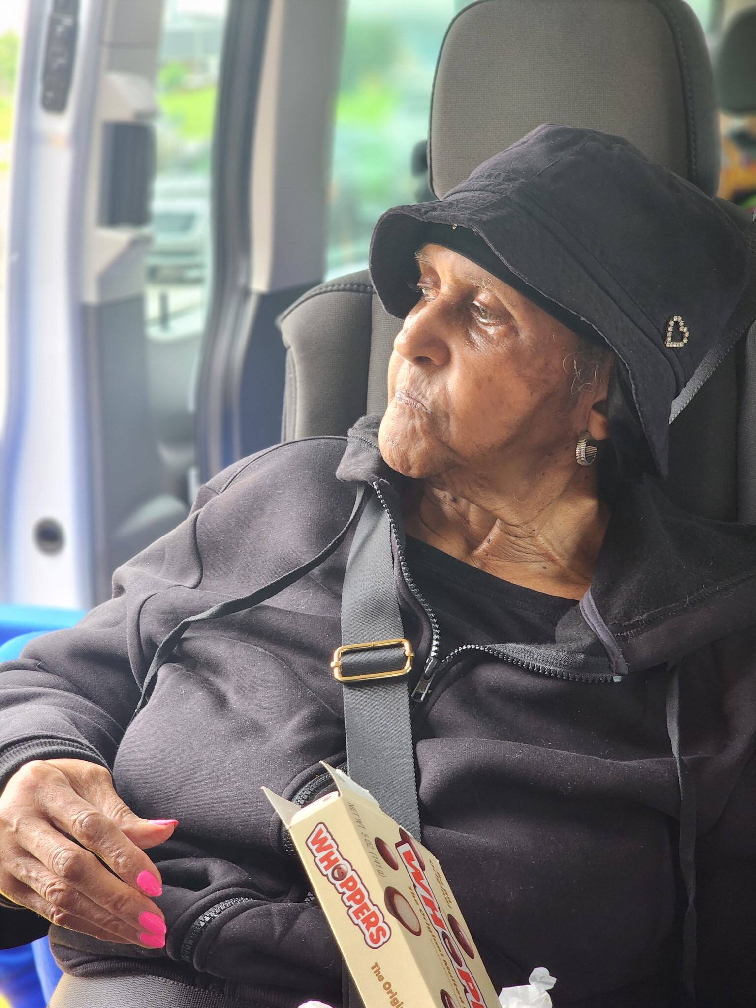 Older woman in a black hoodie wearing a seatbelt sits in a vehicle, holding a Whoppers candy box and looking out the window.