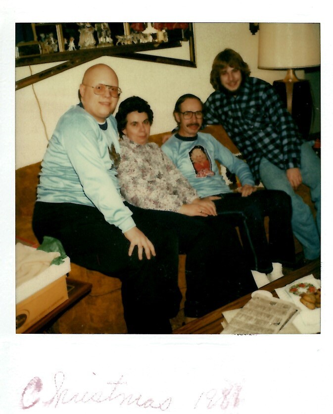Four adults sit close together on a brown sofa in a living room, posing for a family Christmas photo.