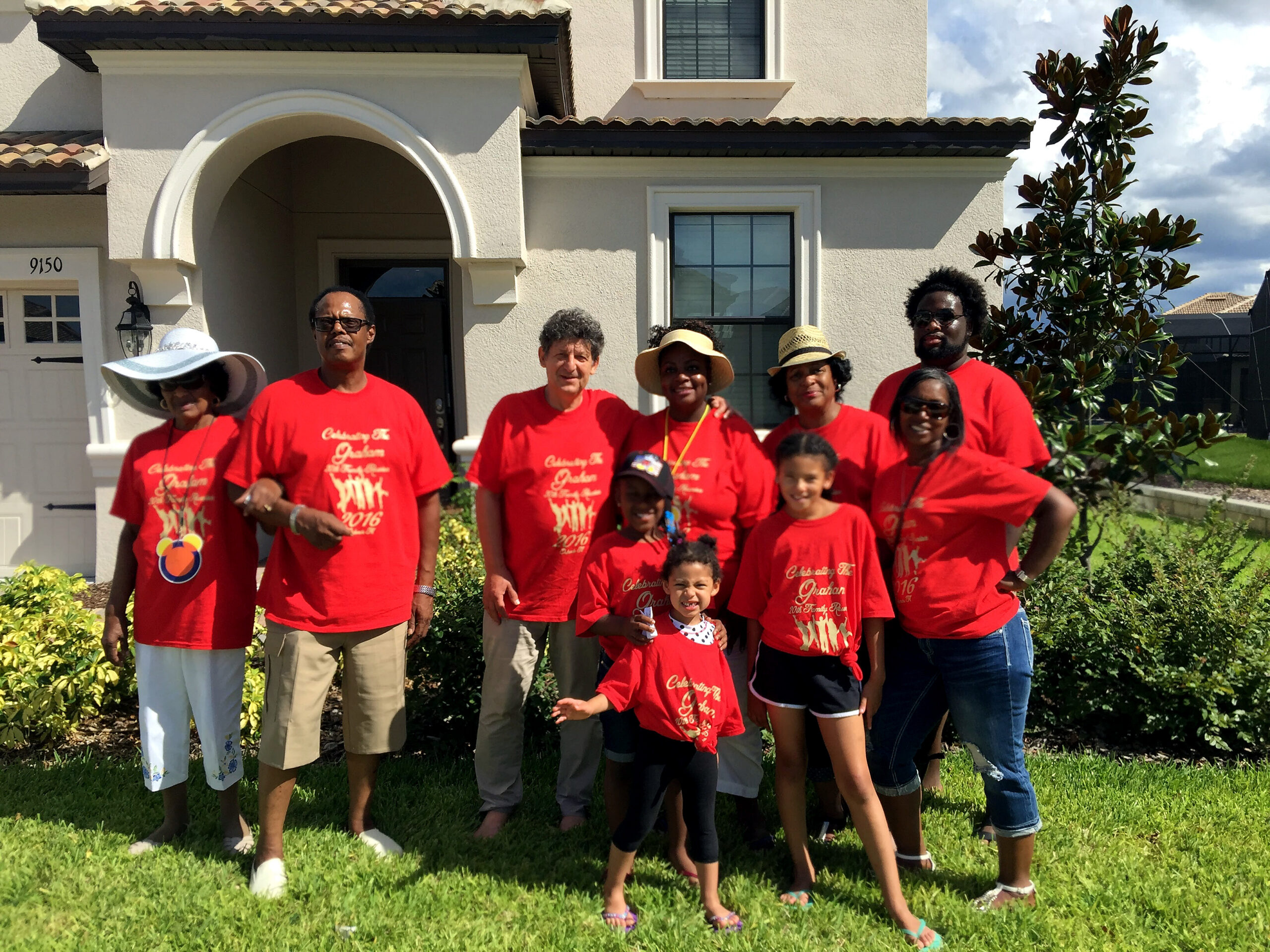 Group of people in red shirts posing on a lawn in front of a beige house with an arched entryway on a sunny day.