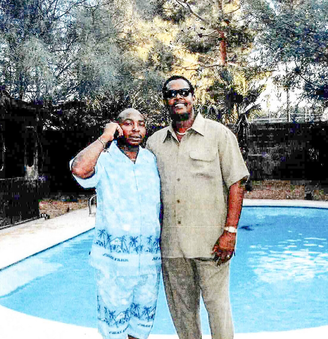 Two adults pose by a backyard pool; man in a beige short-sleeve shirt and sunglasses, woman in a light blue tropical dress.