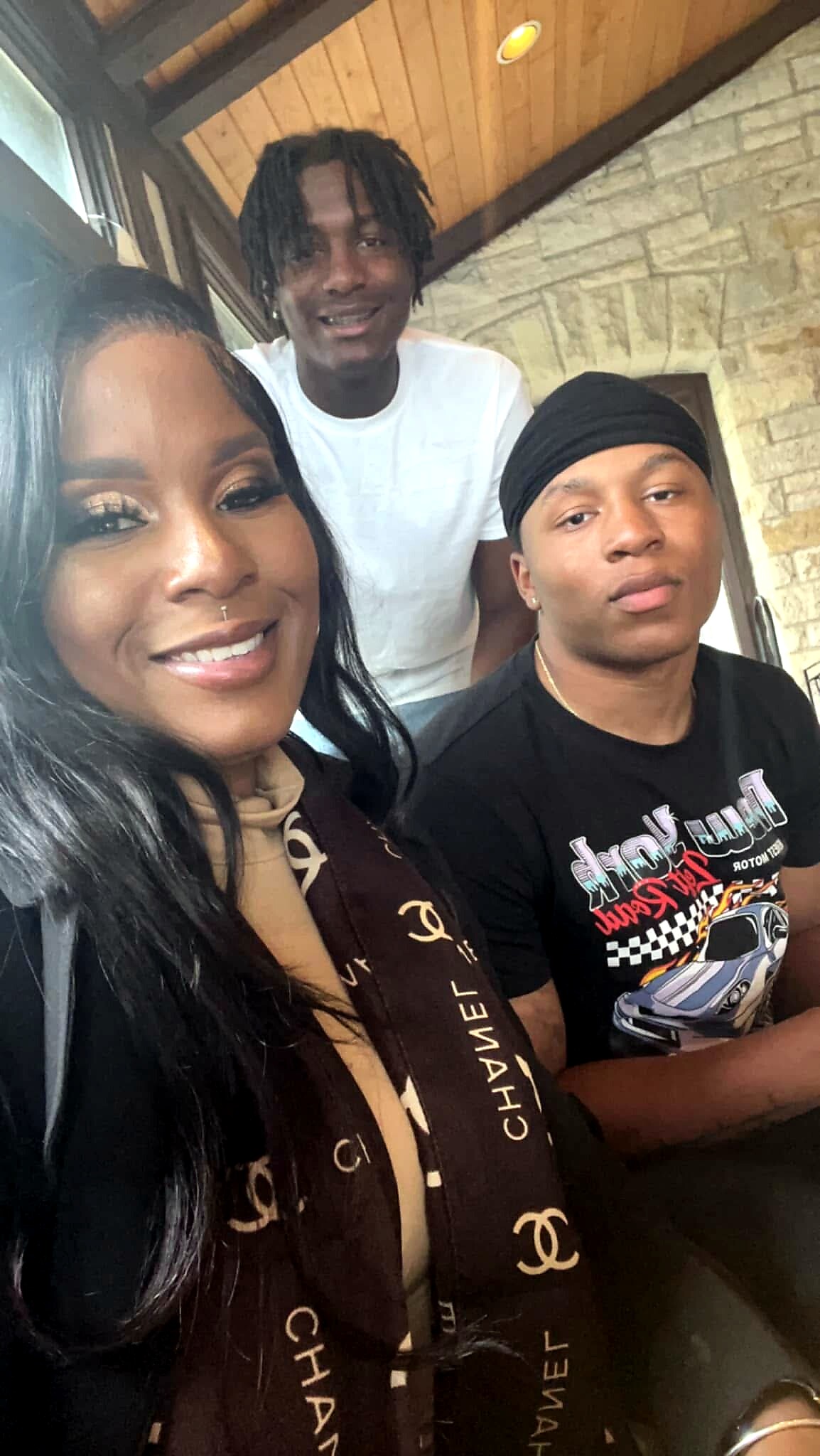Three people indoors posing for a group selfie: a woman in front left smiling, with two men behind against a stone wall and wooden ceiling in the background.