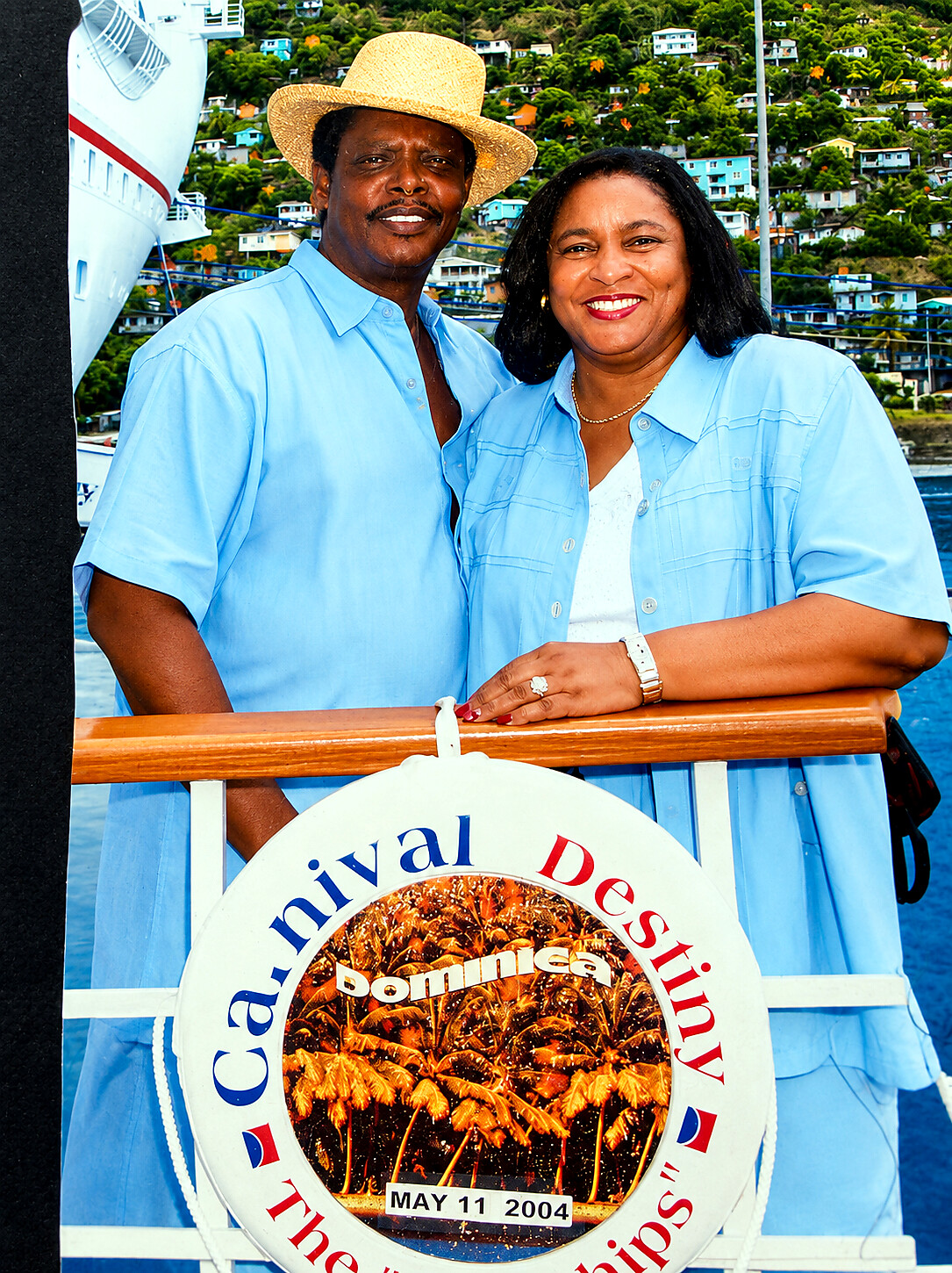 Smiling couple in light-blue shirts on a cruise ship deck, leaning on railing with a circular sign featuring palm trees and the date May 11, 2004 (Dominica cruise).