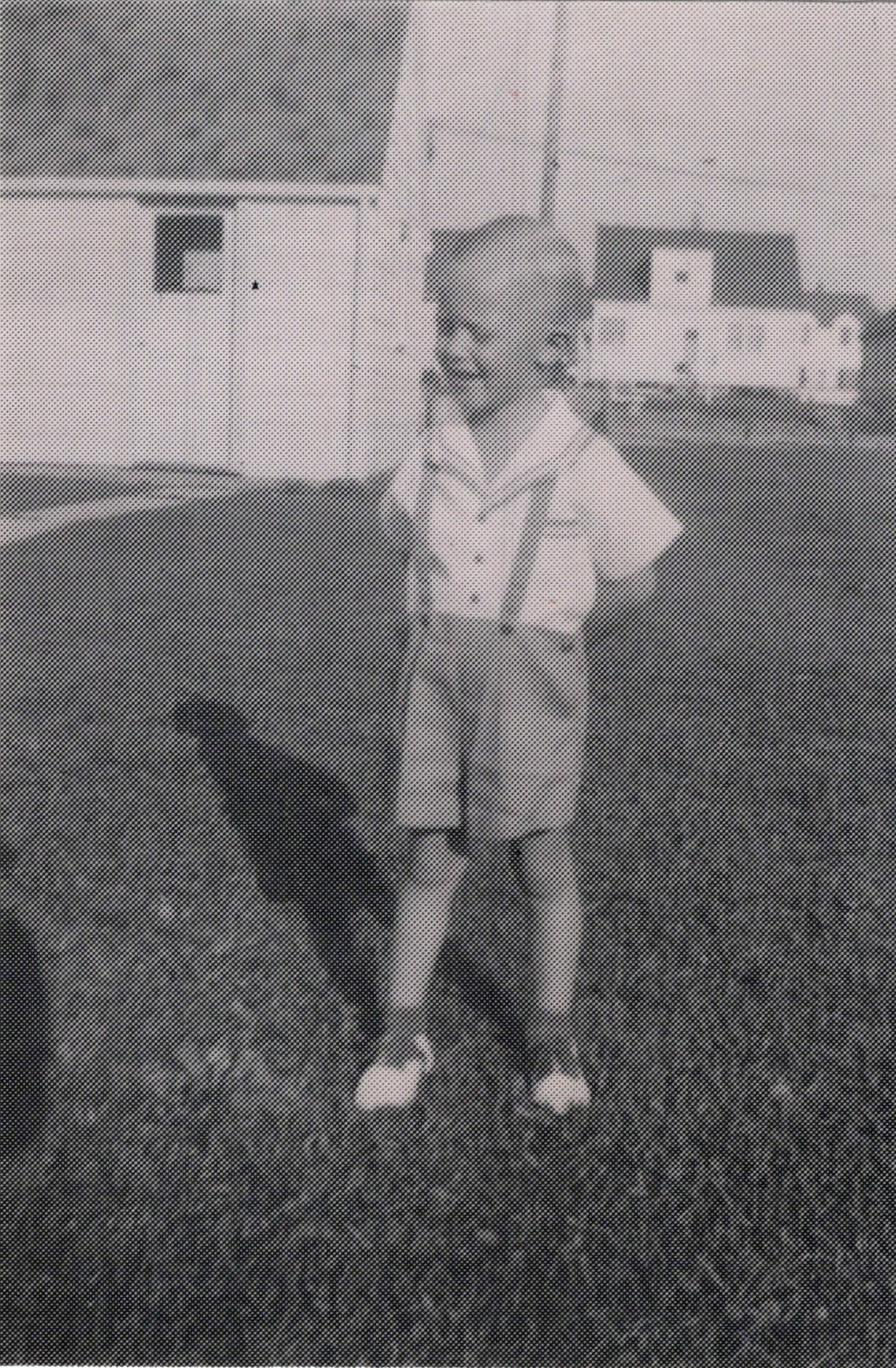 Child in blazer and shorts standing in a grassy yard with a building behind him.