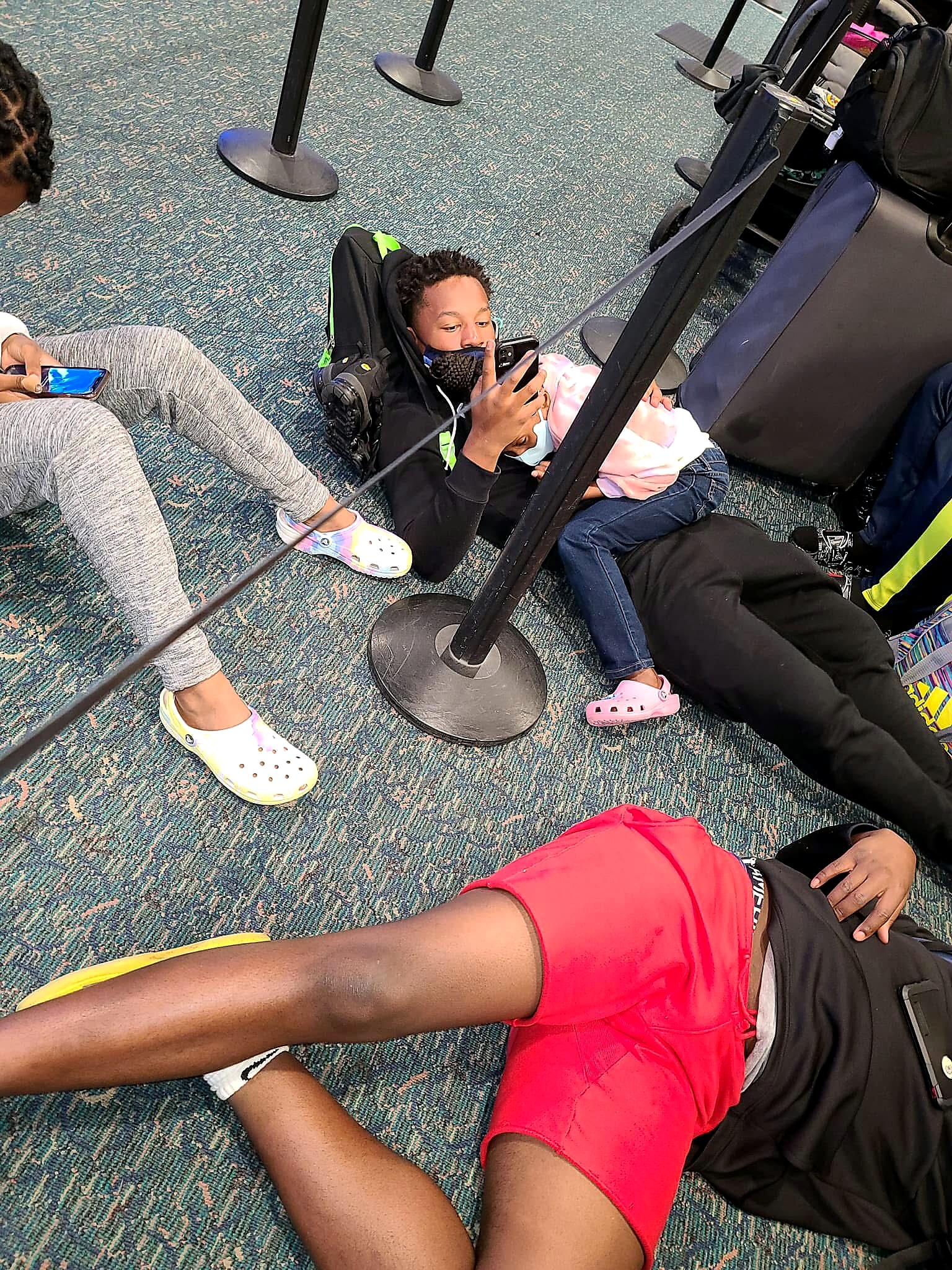Child wearing a mask, reclining against a backpack and scrolling on a phone in an airport-style waiting area.
