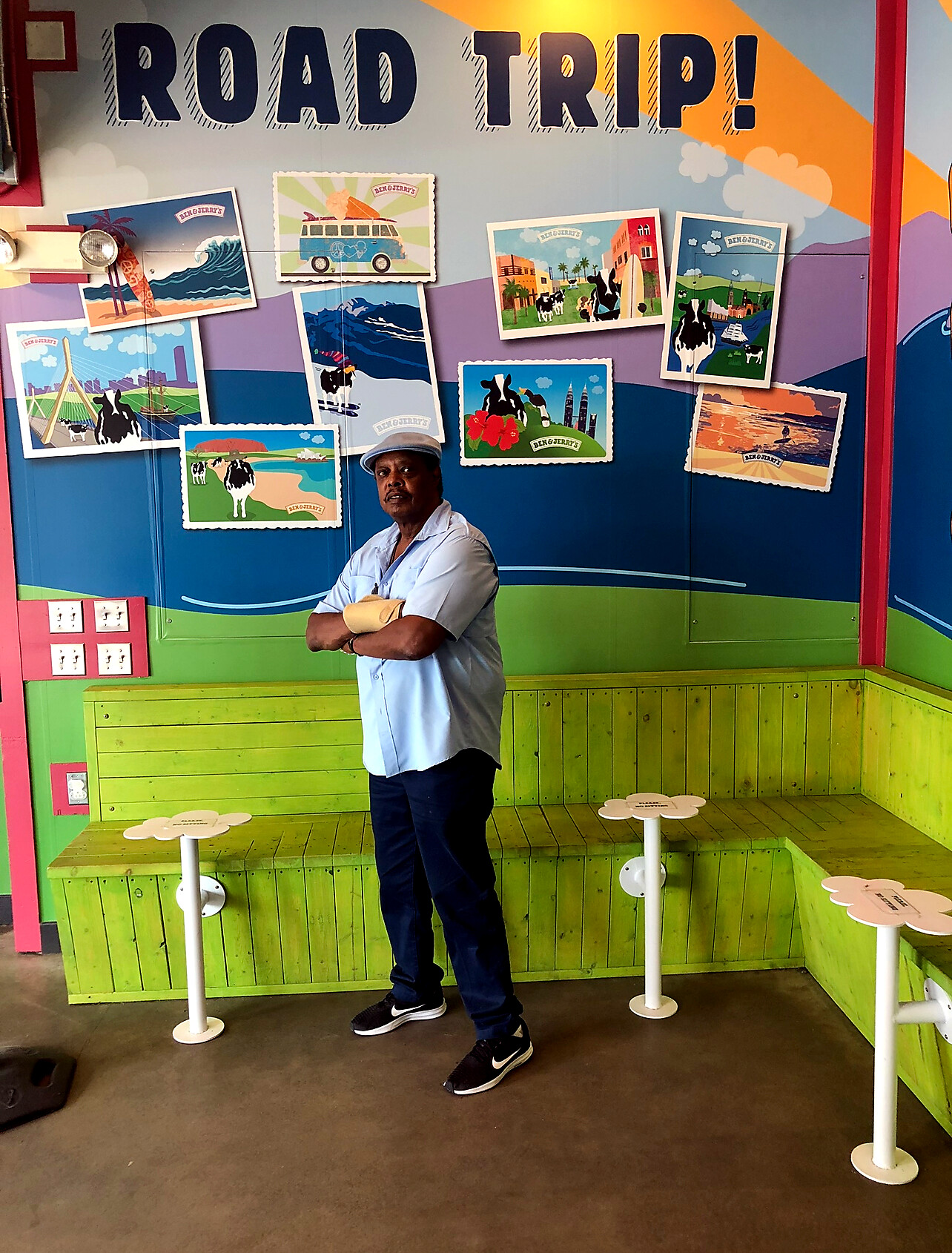 Man in a light blue shirt and cap stands with arms crossed in front of a colorful Ben & Jerry's 'Road Trip' mural with travel posters on the wall.