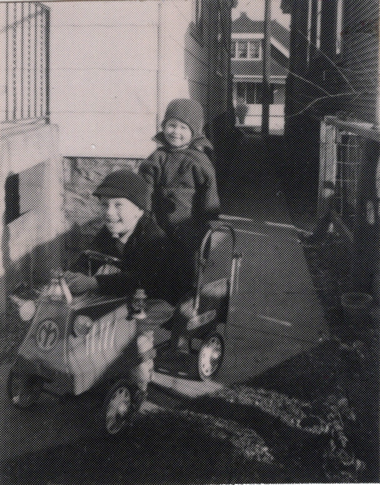 Two young children in winter coats and hats stand beside a baby carriage on a steps outside a house.