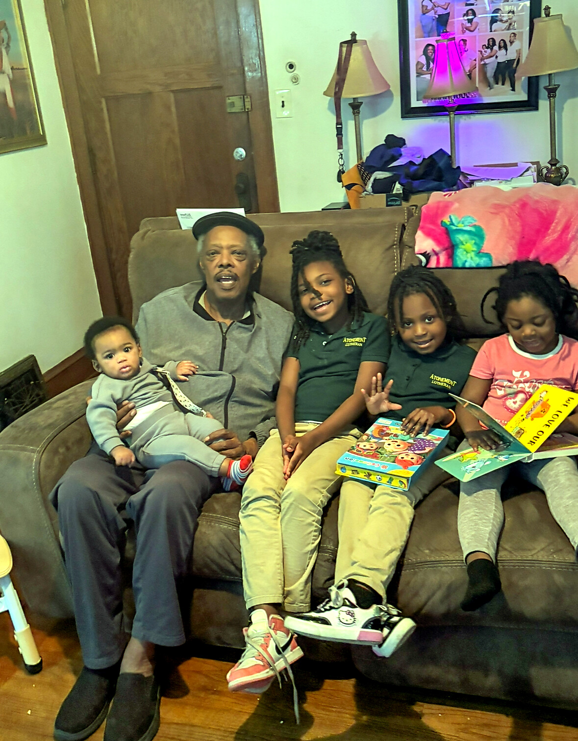 An older man wearing a cap sits on a brown sofa with a baby on his lap, while three children sit beside him reading colorful books in a living room.