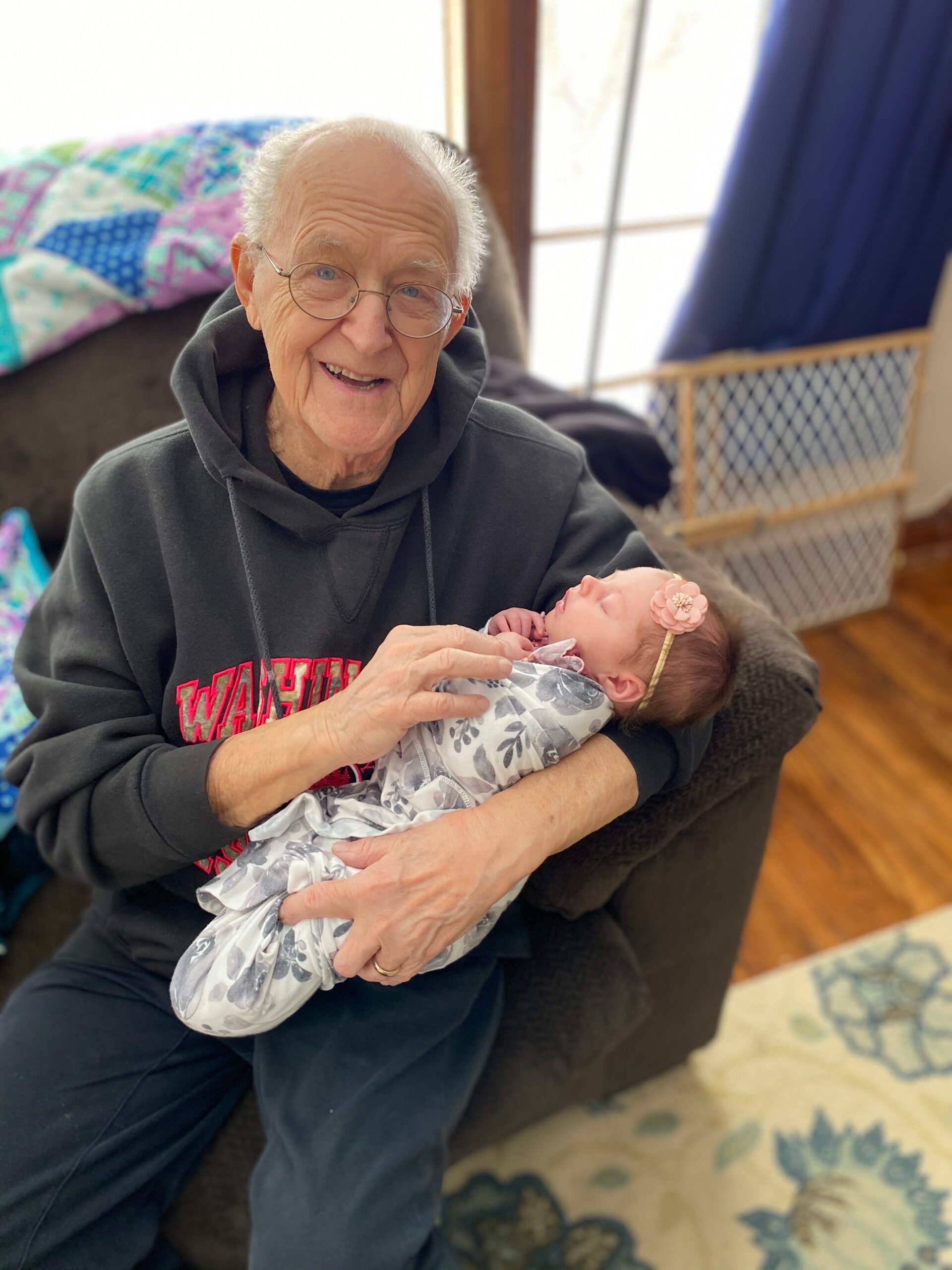 Smiling elderly woman wearing glasses cradling a newborn baby wrapped in a gray floral blanket.
