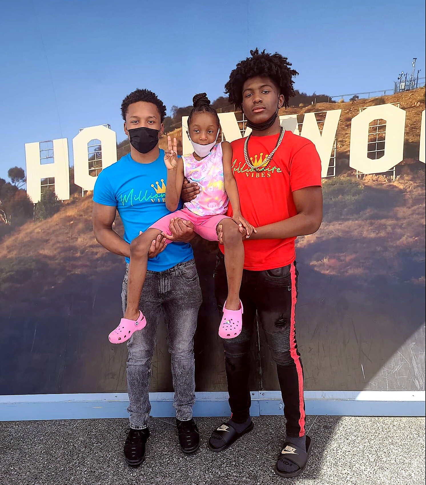 Three people pose together in front of a Hollywood backdrop; a child sits between two men wearing bright shirts, all casual and smiling (some mask worn).