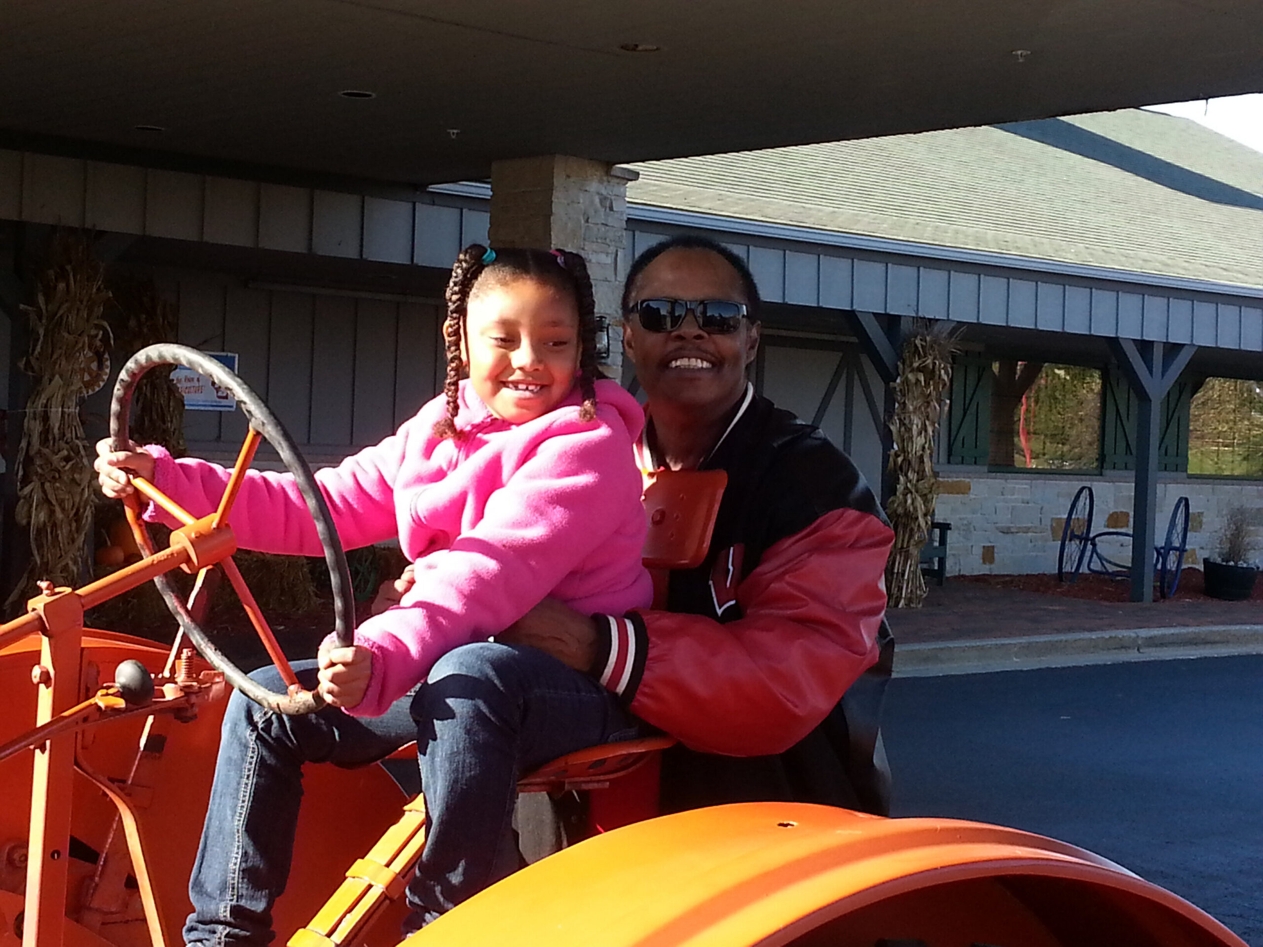 Smiling man and young girl in a pink jacket ride an orange amusement vehicle outdoors, facing the camera.