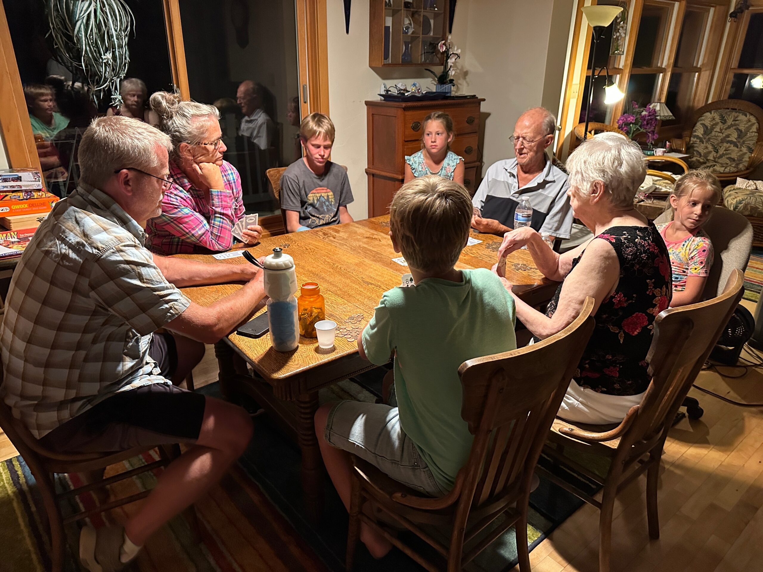 Family group around a wooden dining table playing a card/board game indoors, with coins and cups on the table.