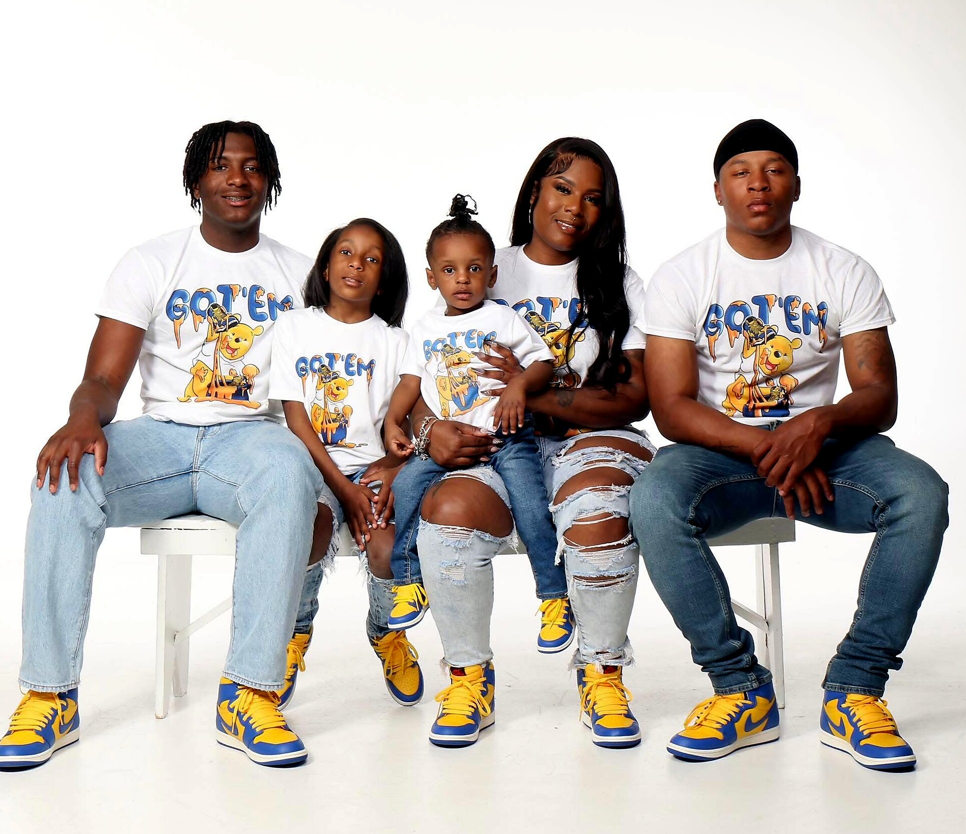 Family of five posing in matching white shirts with a lion graphic, seated on a white bench in a plain studio setting.